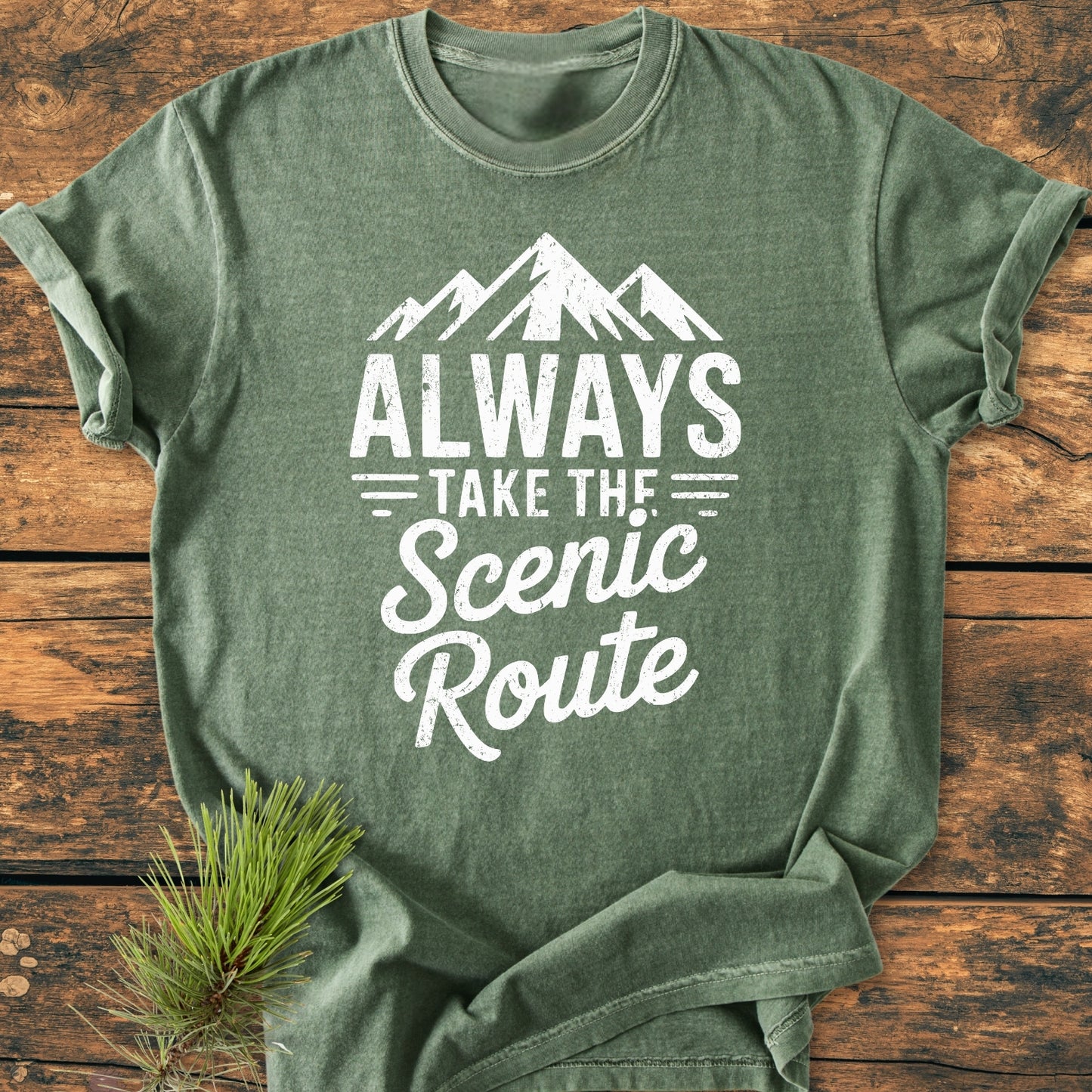 Always Take The Scenic Route - Vintage Wash Tee displayed on wood, featuring a white mountain graphic and text. Includes a pine bough accent for a nature-inspired theme.