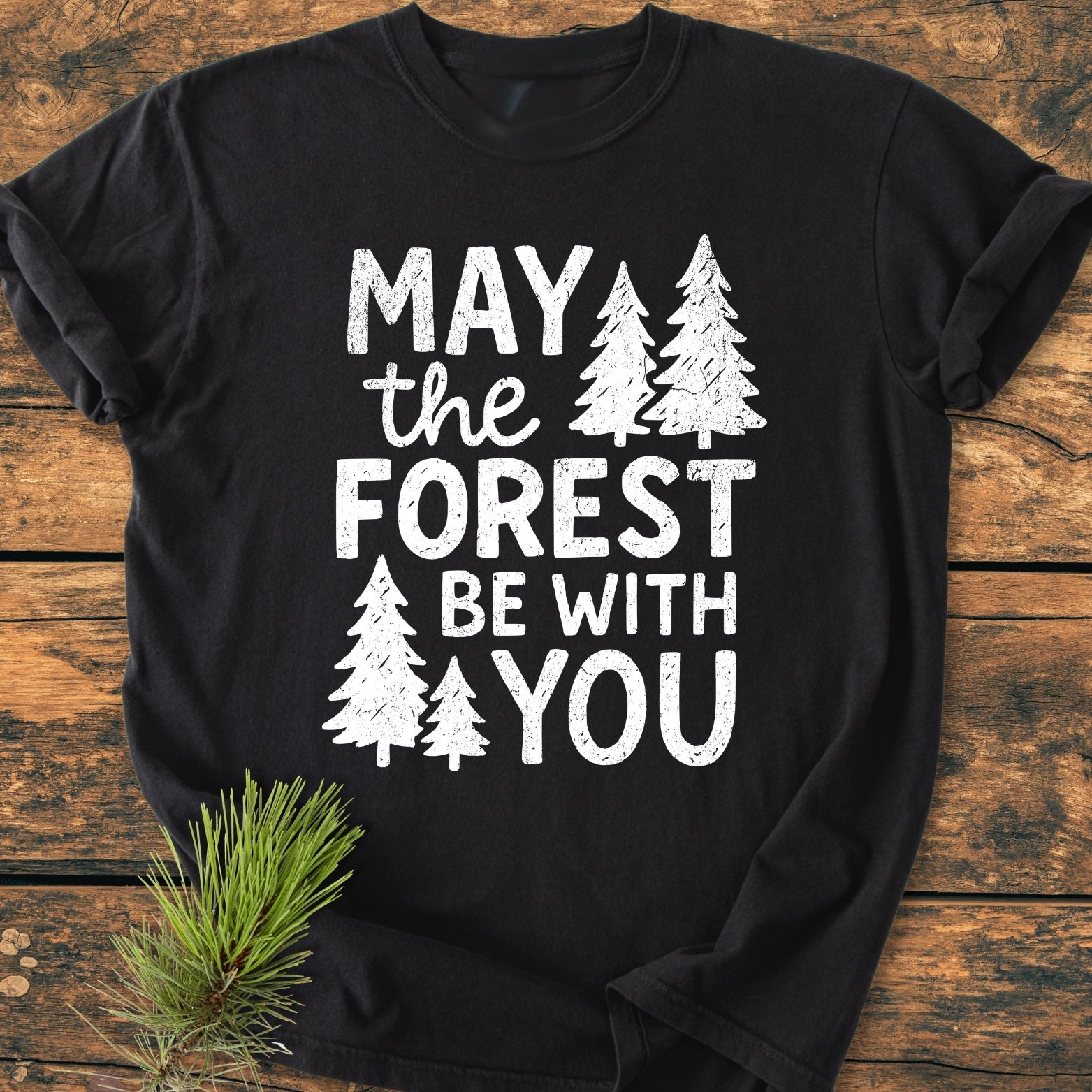 May The Forest Be With You - Vintage Wash Tee displayed on a wooden surface, featuring a white, distressed print with pine tree silhouettes, complemented by a small pine branch beside it.