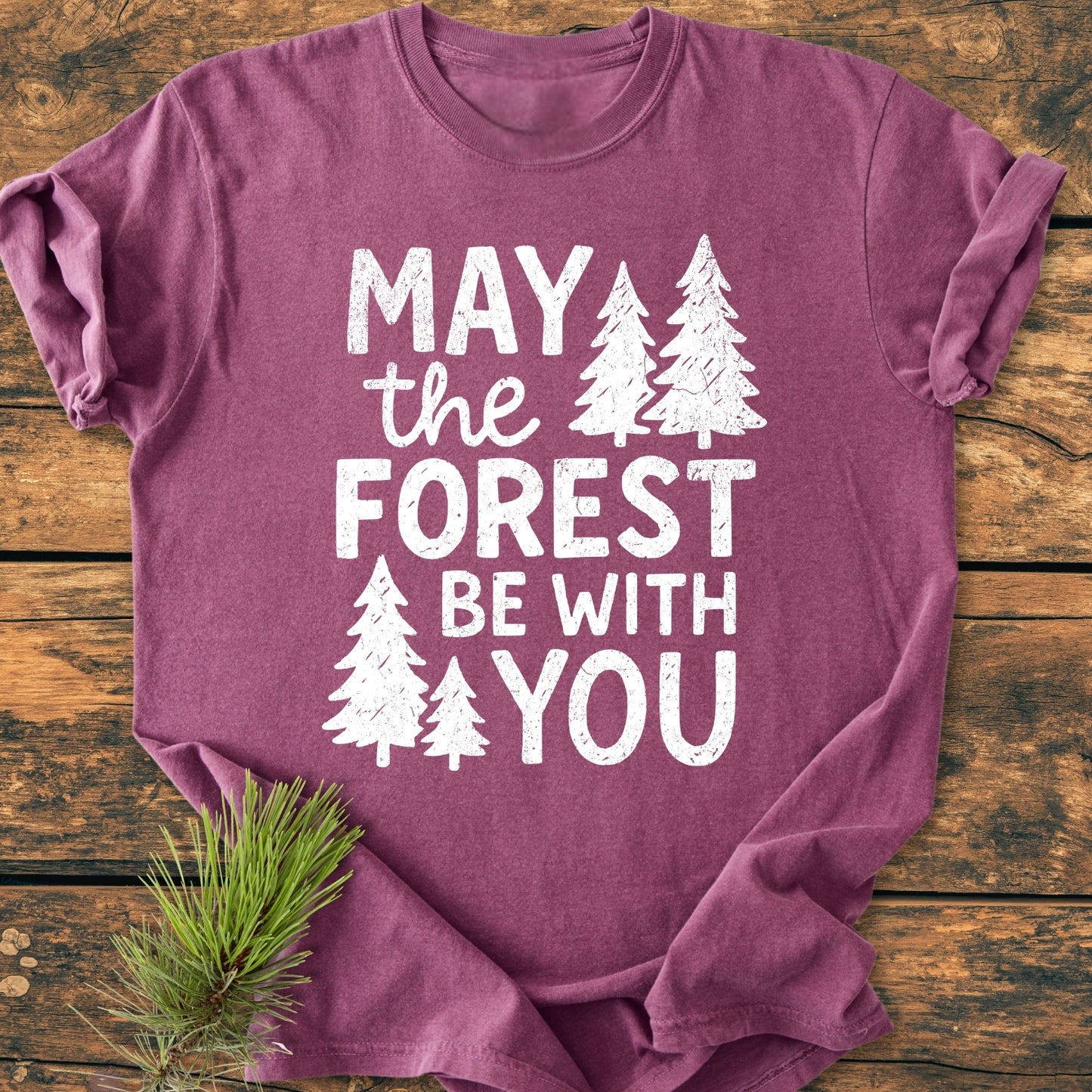 May The Forest Be With You - Vintage Wash Tee features a heather-purple color with white pine-tree print, laid flat on a wooden surface, complemented by a small pine needle sprig.