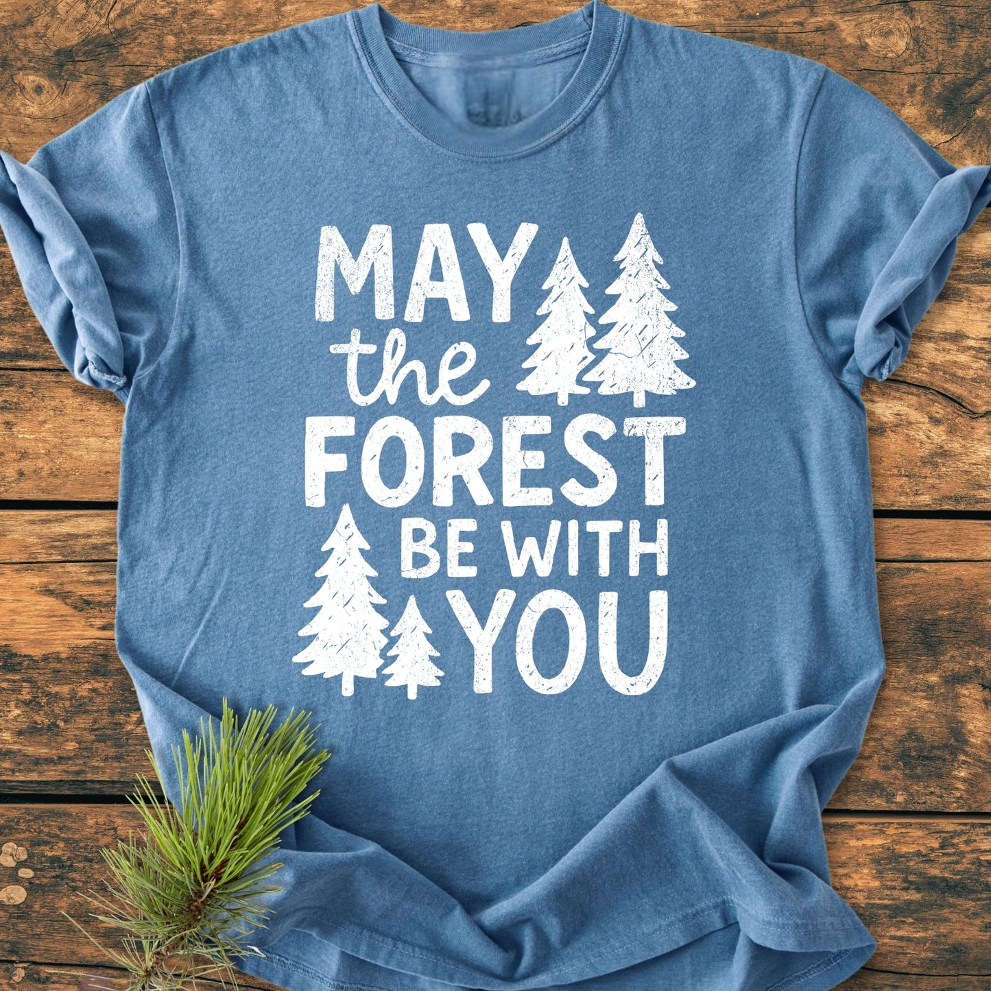 Heather-blue May The Forest Be With You - Vintage Wash Tee featuring distressed white lettering and pine-tree silhouettes, displayed on wood, with a pine needle sprig beside it.