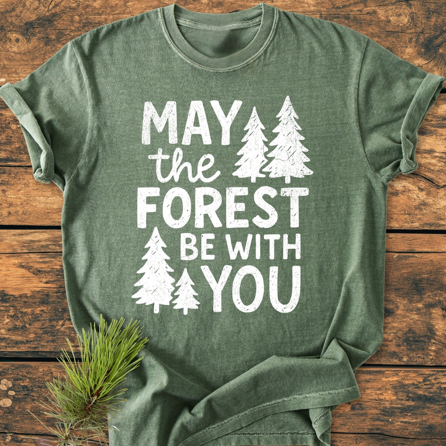 Green May The Forest Be With You - Vintage Wash Tee on rustic wood, featuring white text with pine-tree silhouettes. Accompanied by a pine needle sprig, highlighting its nature-inspired theme.