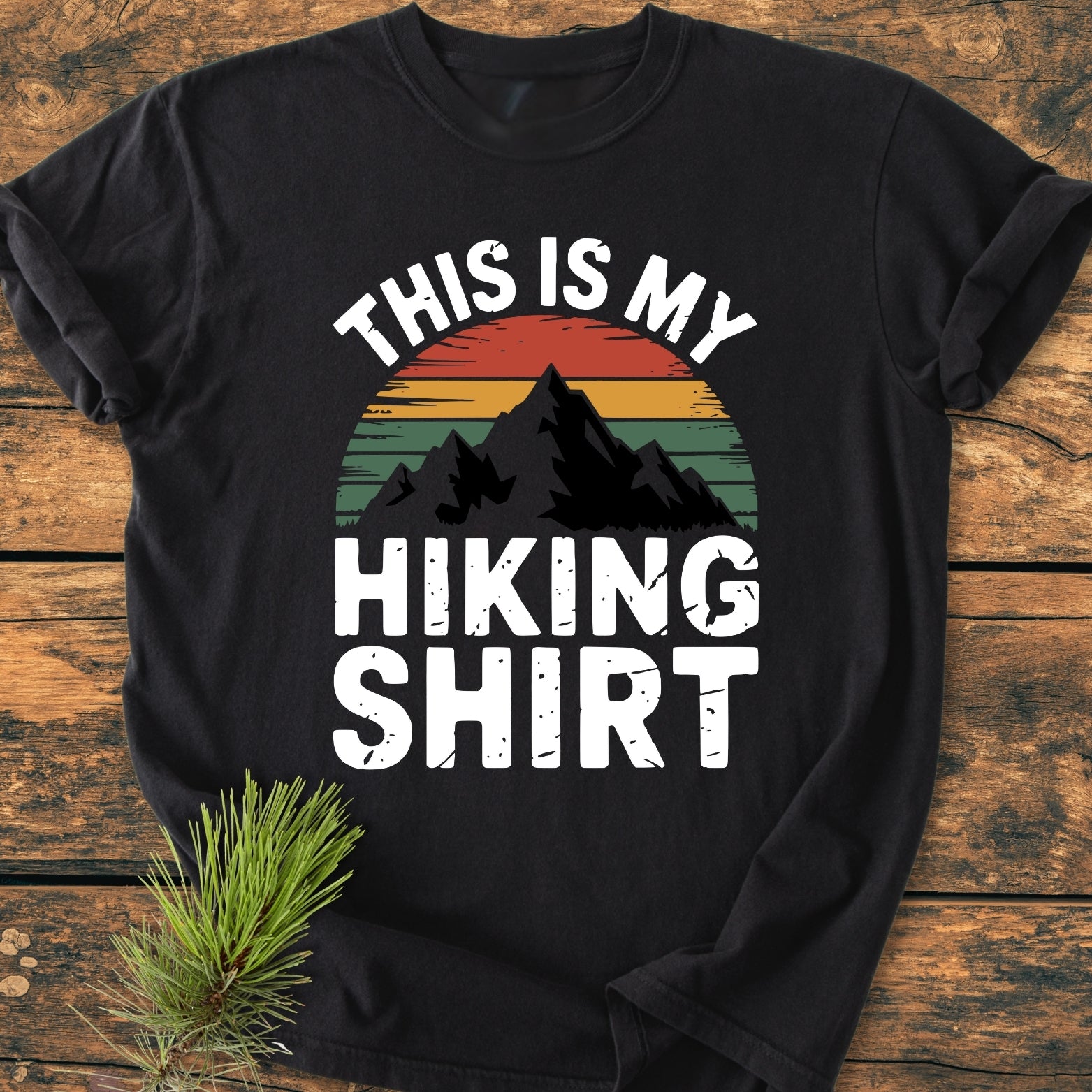 This Is My Hiking Shirt - Vintage Wash Tee: Black T-shirt featuring a mountain silhouette with a retro-striped sunset design and bold white text. Laid on wood, pine sprig accentuates the scene.