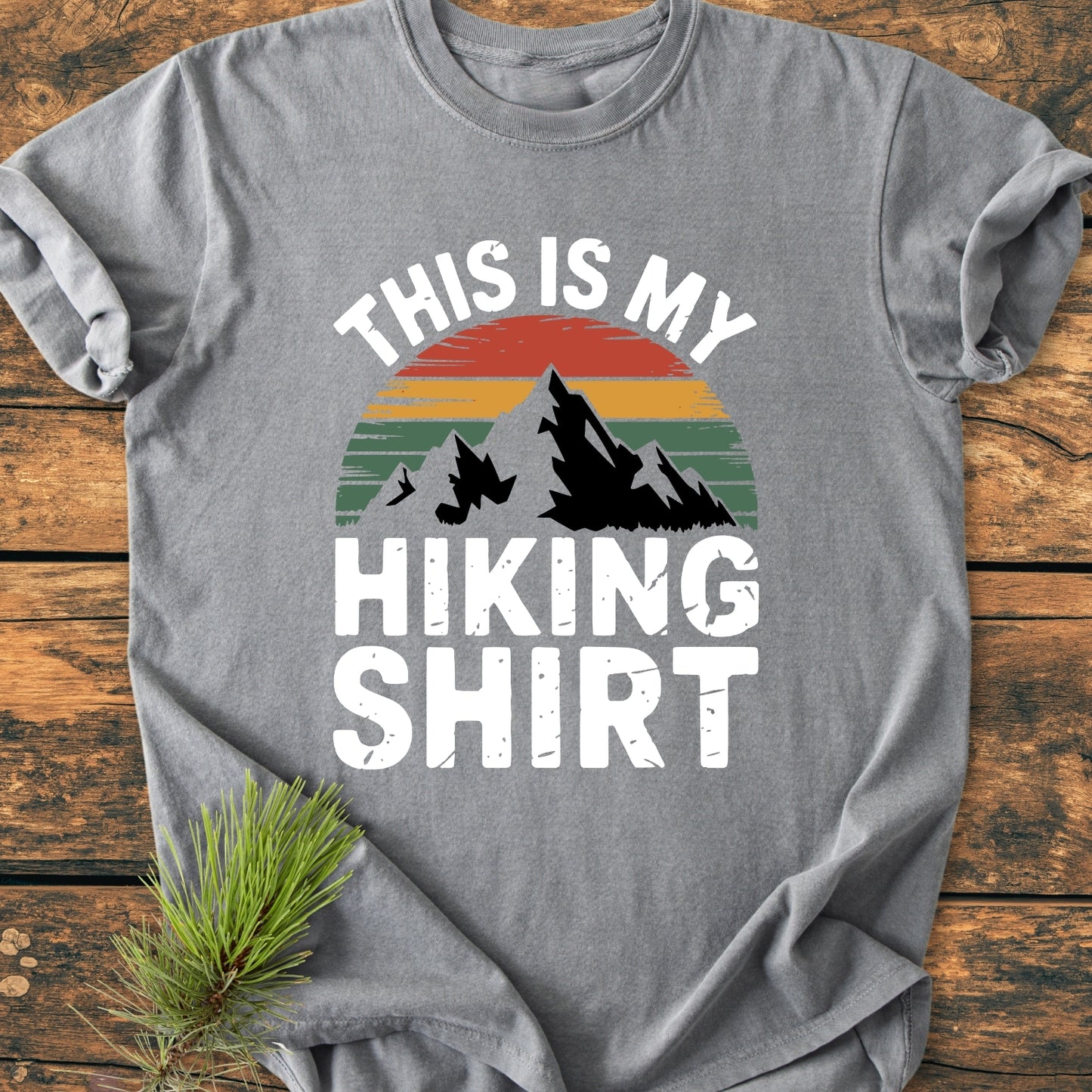 This Is My Hiking Shirt - Vintage Wash Tee features a mountain graphic with a sunset design. Laid on wood, the heather-gray tee includes bold lettering and a pine sprig accentuating its outdoor theme.