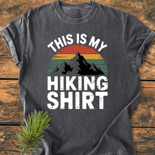 This Is My Hiking Shirt - Vintage Wash Tee features a mountain silhouette with a sunset graphic, laid on a wooden surface. The shirt, made of soft garment-dyed cotton, offers a relaxed fit.