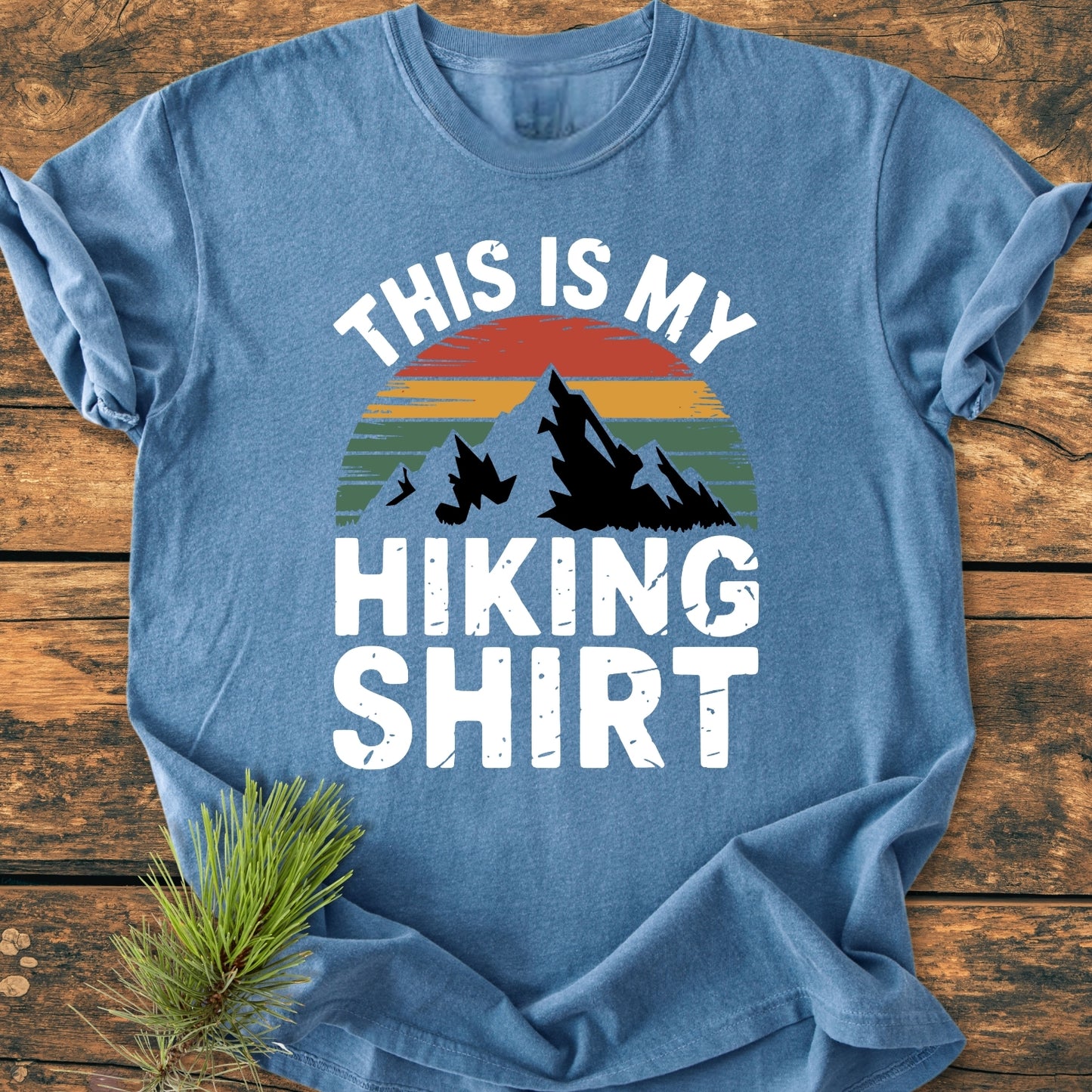 This Is My Hiking Shirt - Vintage Wash Tee features a mountain and sunset graphic on a heather-blue T-shirt, laid on a wooden surface with a pine branch accent for a rustic touch.