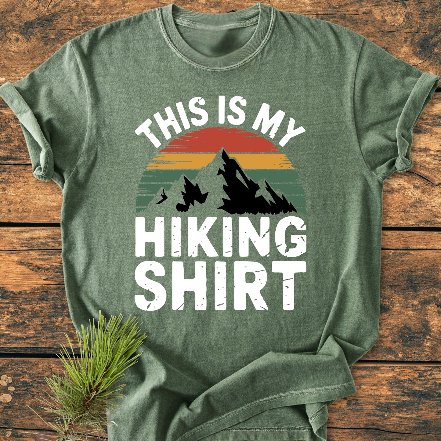 The This Is My Hiking Shirt - Vintage Wash Tee features a heather-green color with a sunset/mountain graphic and bold white text, complemented by a rustic pine branch.