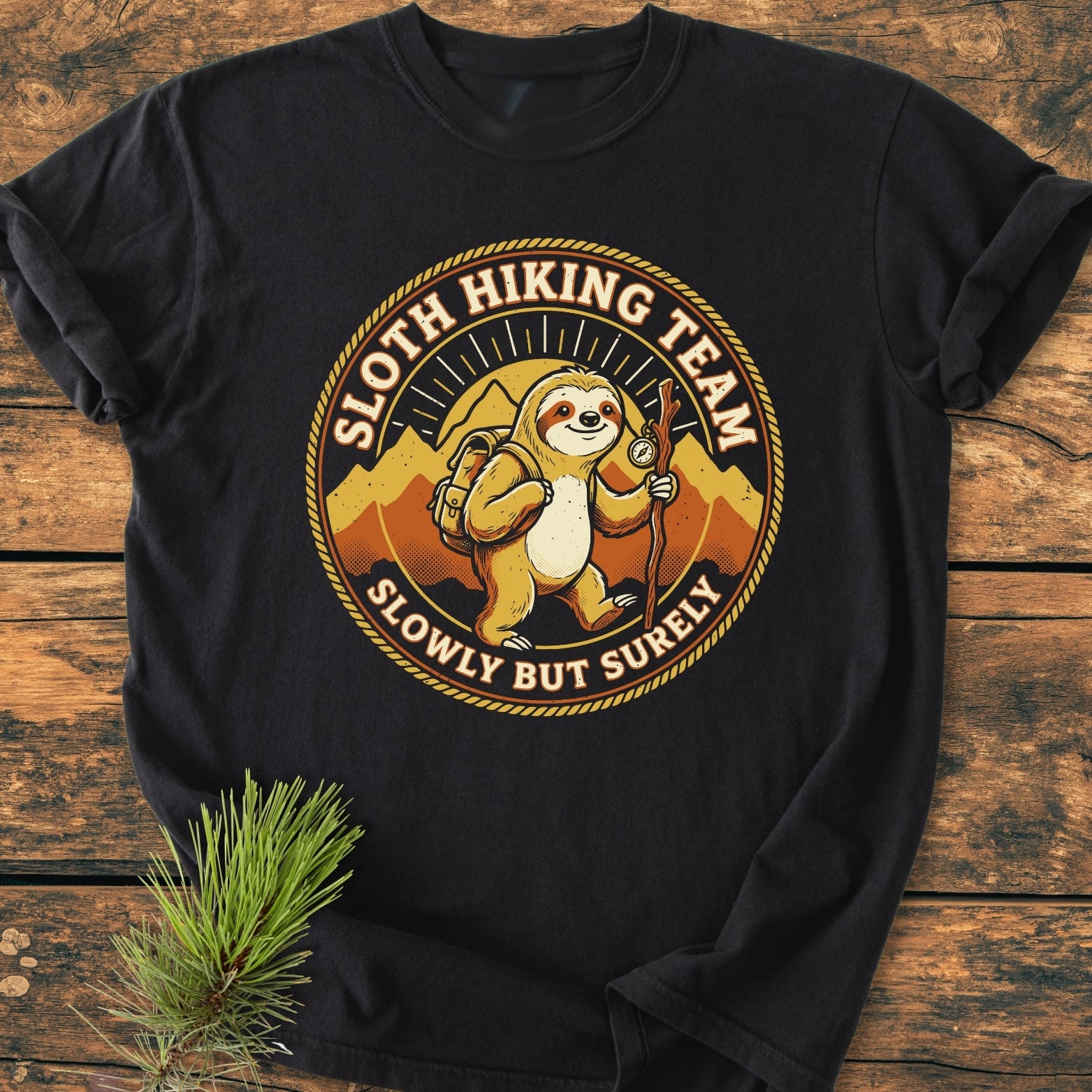Sloth Hiking Team - Vintage Wash Tee featuring a cartoon sloth with a backpack and walking stick, surrounded by mountains and sunburst, laid on a wooden surface with a pine sprig.
