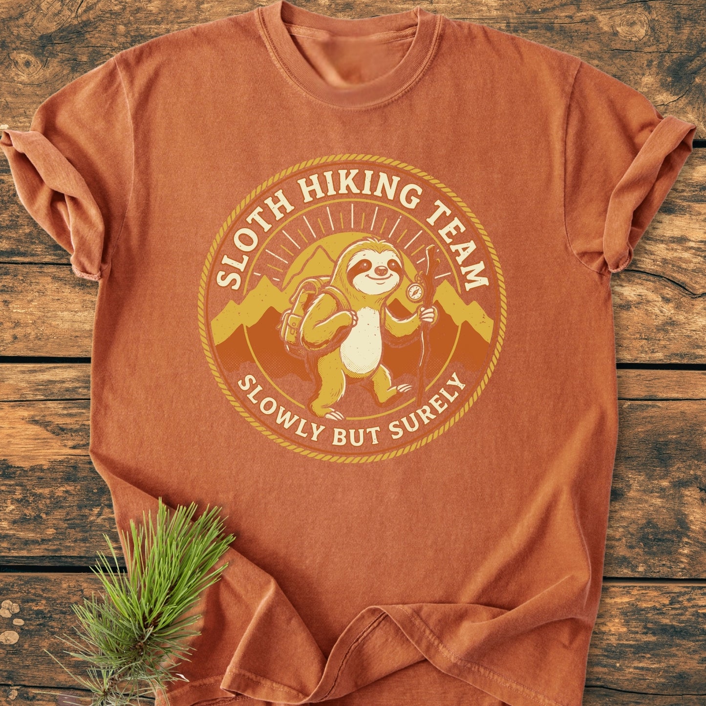 Sloth Hiking Team - Vintage Wash Tee featuring a cartoon sloth with hiking gear and mountains, laid flat on wood. Includes slogan SLOWLY BUT SURELY. Beside it, a pine sprig enhances the outdoor theme.