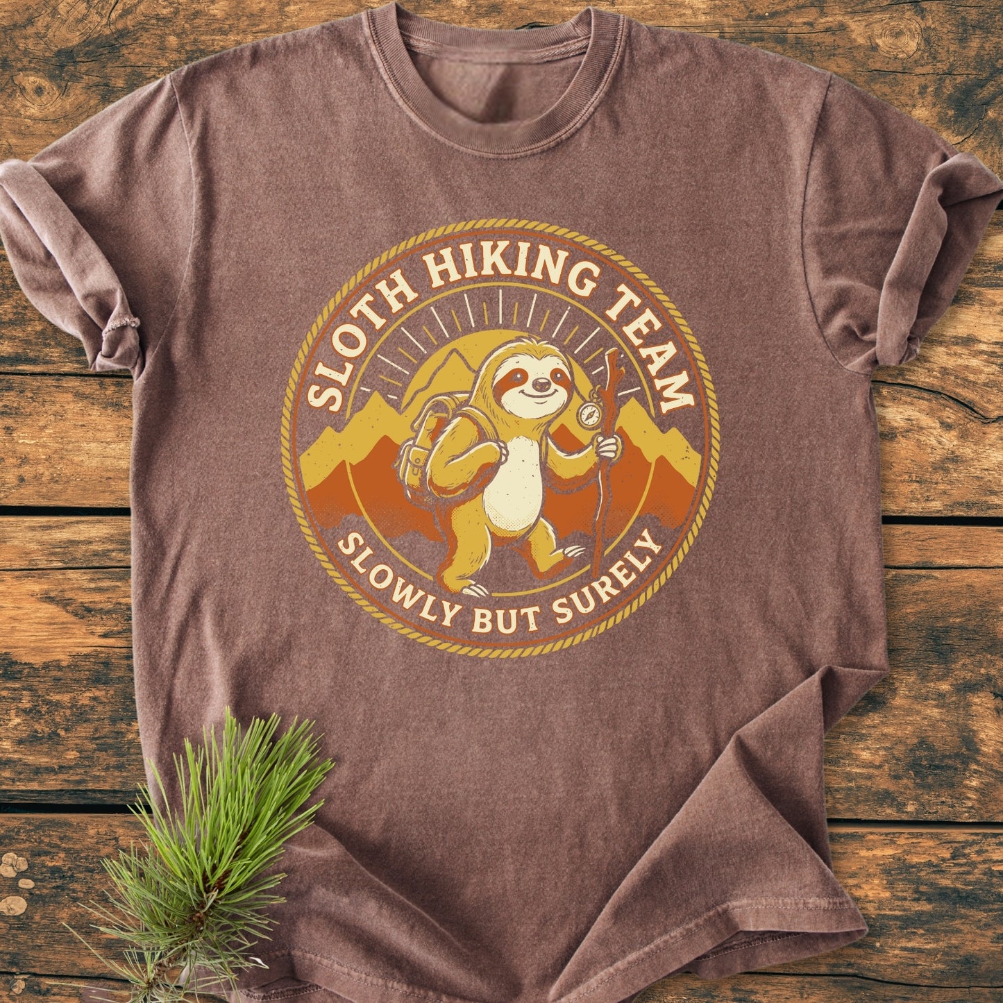 Sloth Hiking Team - Vintage Wash Tee featuring a cartoon sloth with hiking gear and mountains. The design reads “SLOTH HIKING TEAM, SLOWLY BUT SURELY.” Laid flat with a pine sprig beside it.