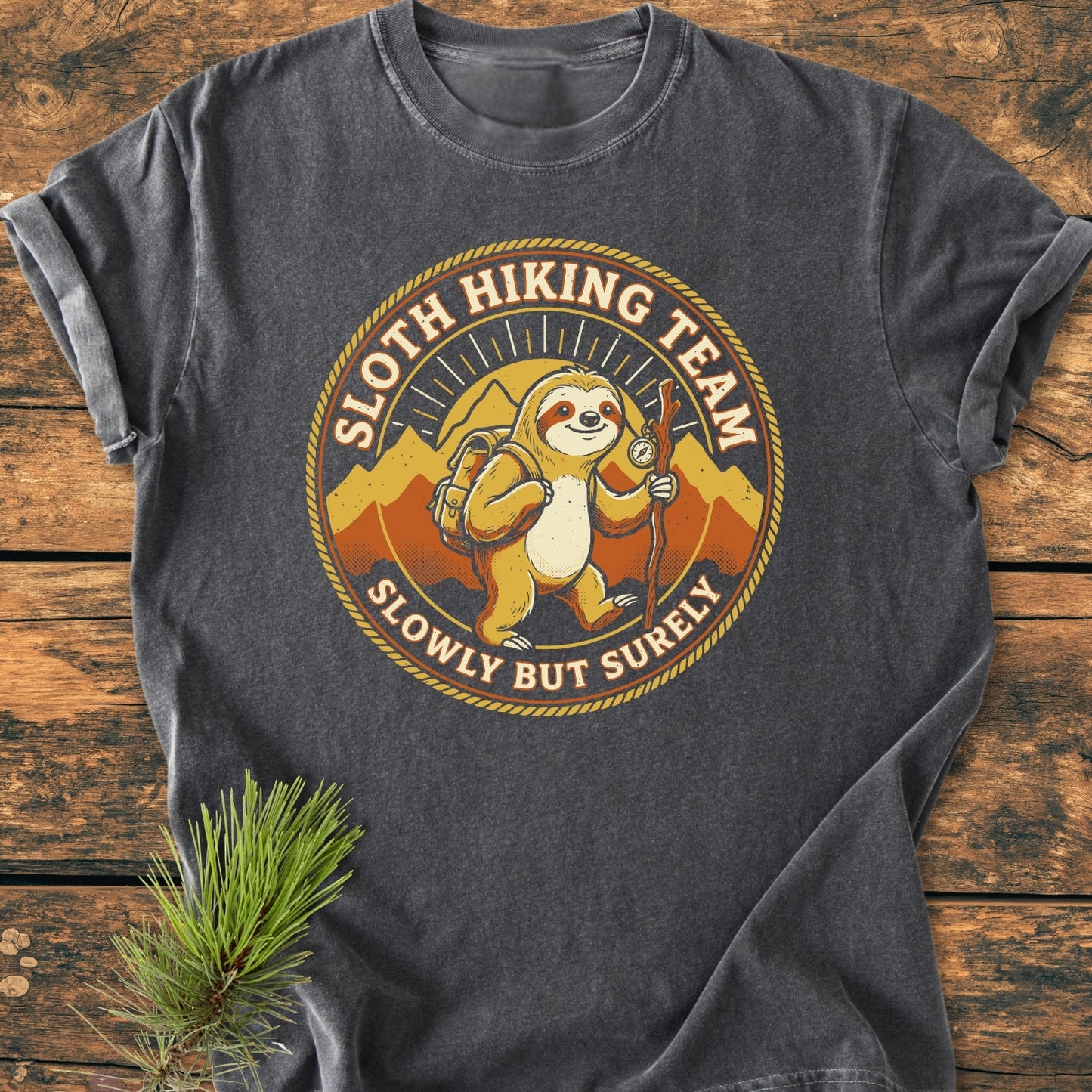 Sloth Hiking Team - Vintage Wash Tee features a cartoon sloth emblem with hiking gear and mountains, laid flat on a wooden surface, showcasing its relaxed fit and durable, soft-washed cotton fabric.