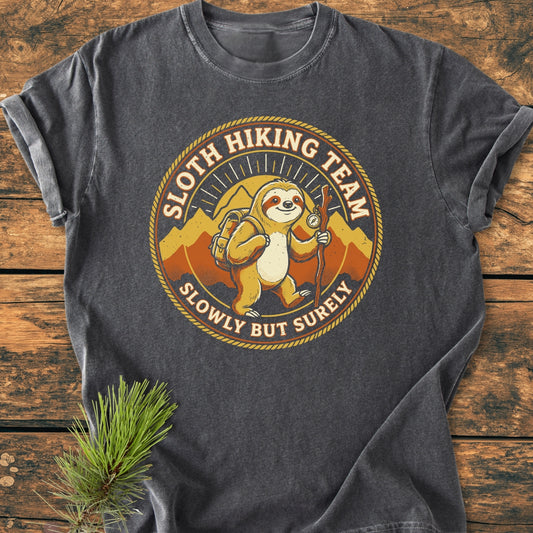 Sloth Hiking Team - Vintage Wash Tee features a cartoon sloth emblem with hiking gear and mountains, laid flat on a wooden surface, showcasing its relaxed fit and durable, soft-washed cotton fabric.