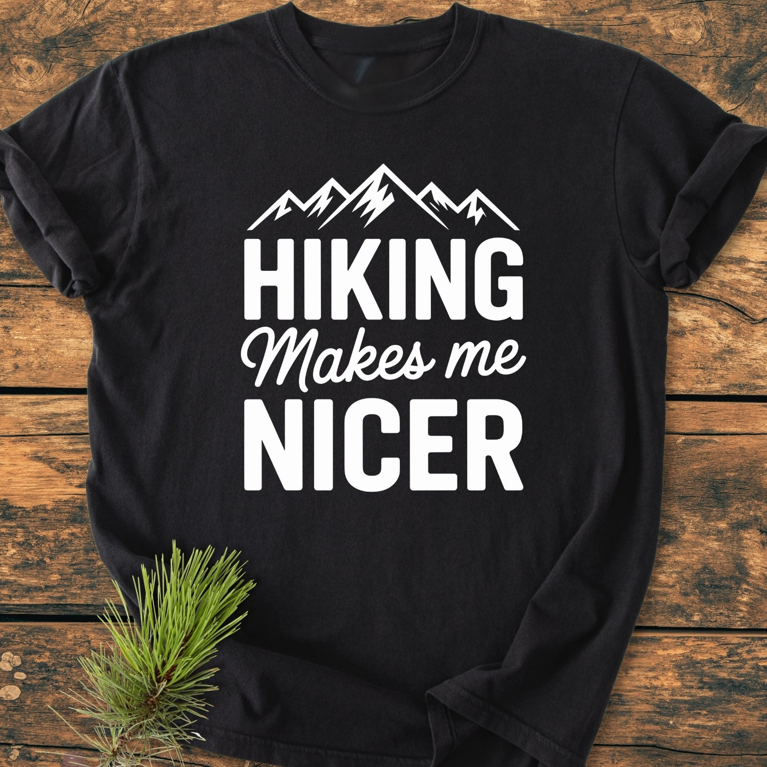 Hiking Makes Me Nicer - Vintage Wash Tee: Black tee with white mountain silhouette and slogan, laid on a wooden surface with a pine sprig accent. Soft-washed, relaxed fit, durable stitching.