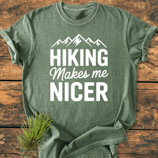 Hiking Makes Me Nicer - Vintage Wash Tee: Forest-green, 100% ring-spun cotton T-shirt with mountain silhouette and slogan, laid on wood; pine needles accentuate its outdoorsy theme.