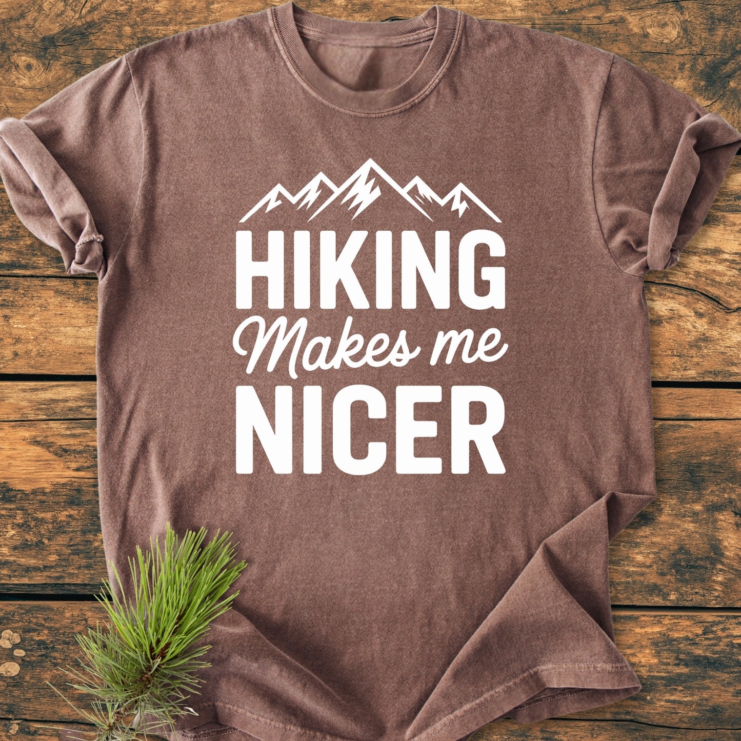 Hiking Makes Me Nicer - Vintage Wash Tee features a mountain graphic and slogan, laid flat on wood with a pine branch, showcasing its garment-dyed, 100% ring-spun cotton, soft and durable design.