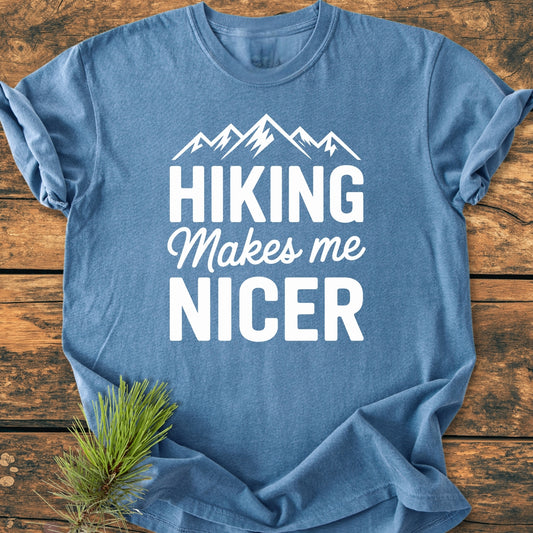 The Hiking Makes Me Nicer - Vintage Wash Tee features a mountain graphic and slogan on a heather-blue T-shirt, laid on wood with pine sprig, emphasizing its relaxed, durable design.