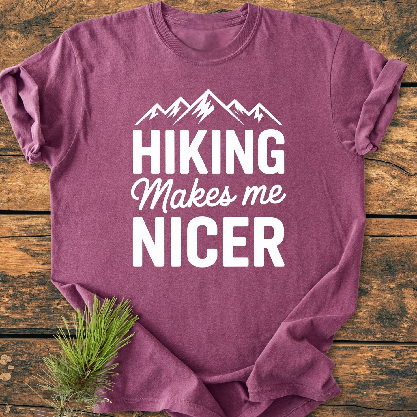 Hiking Makes Me Nicer - Vintage Wash Tee displayed on a wooden surface, featuring a white mountain silhouette and slogan. Accented with a pine sprig, highlighting its outdoorsy theme and relaxed fit.