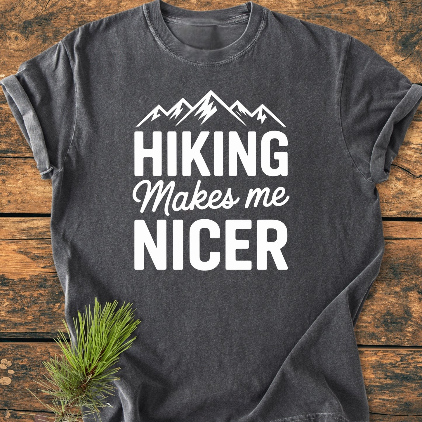 Hiking Makes Me Nicer - Vintage Wash Tee: Dark-grey T-shirt with stylized mountain peaks graphic and slogan, laid on wooden surface, featuring a relaxed fit and soft-washed, garment-dyed fabric.