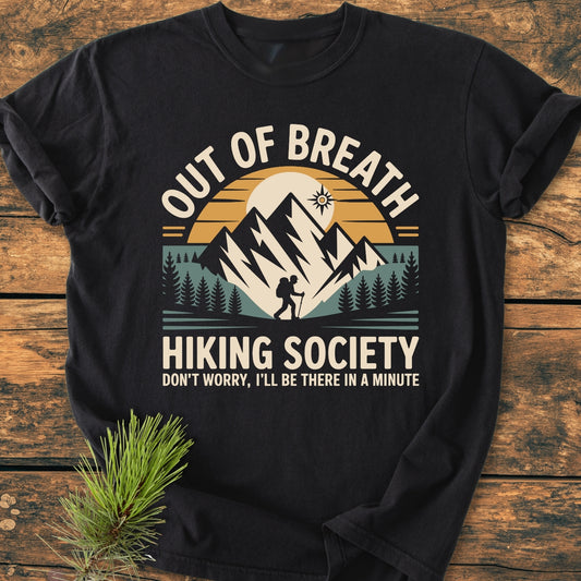 Out Of Breath Hiking Society - Vintage Wash Tee featuring a stylized outdoor scene with mountains, pine trees, and hiker graphic, laid flat on a wooden surface with a nearby pine branch.