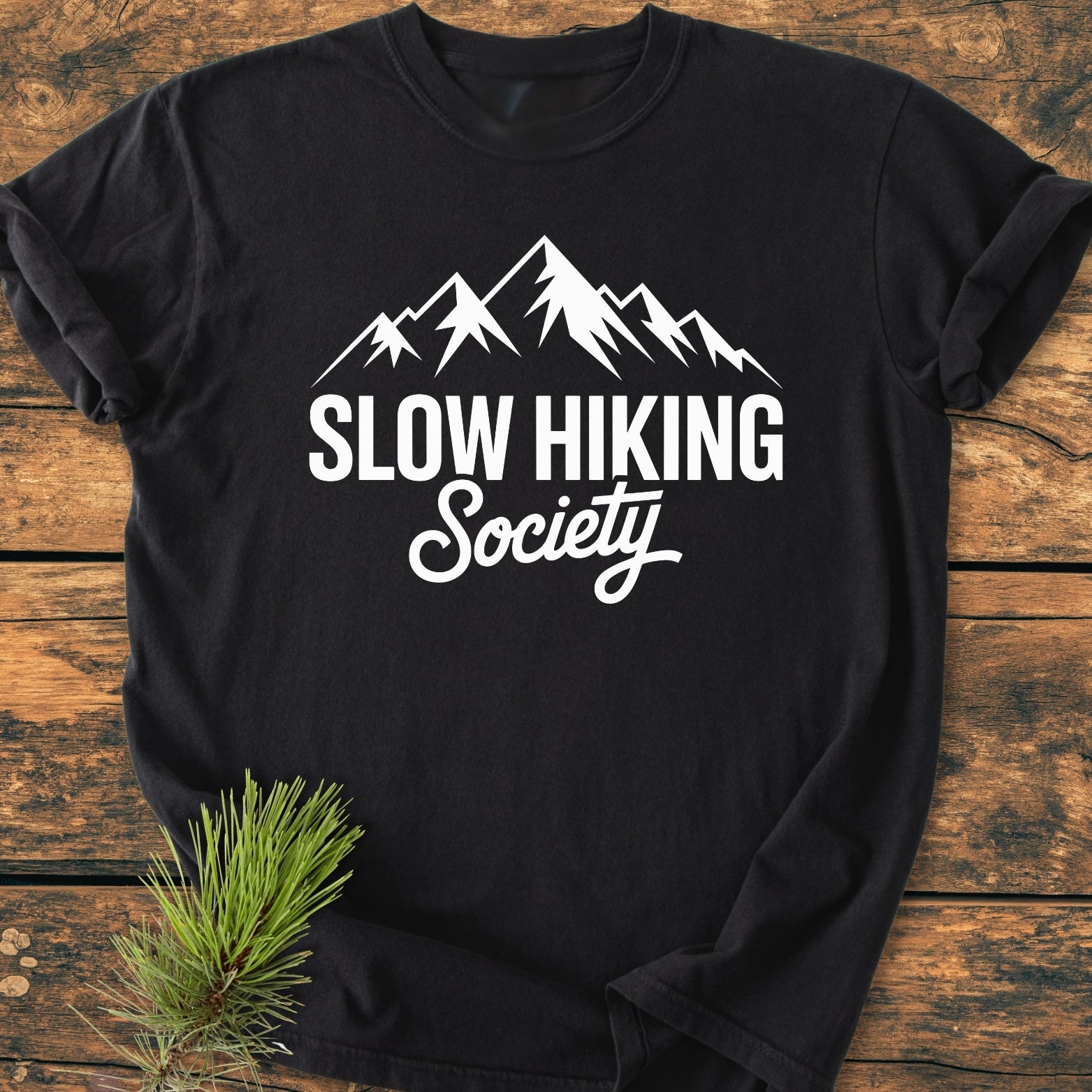 Slow Hiking Society - Vintage Wash Tee features a mountain silhouette and text on a black T-shirt, laid on wood, with a pine sprig accentuating its outdoorsy theme.