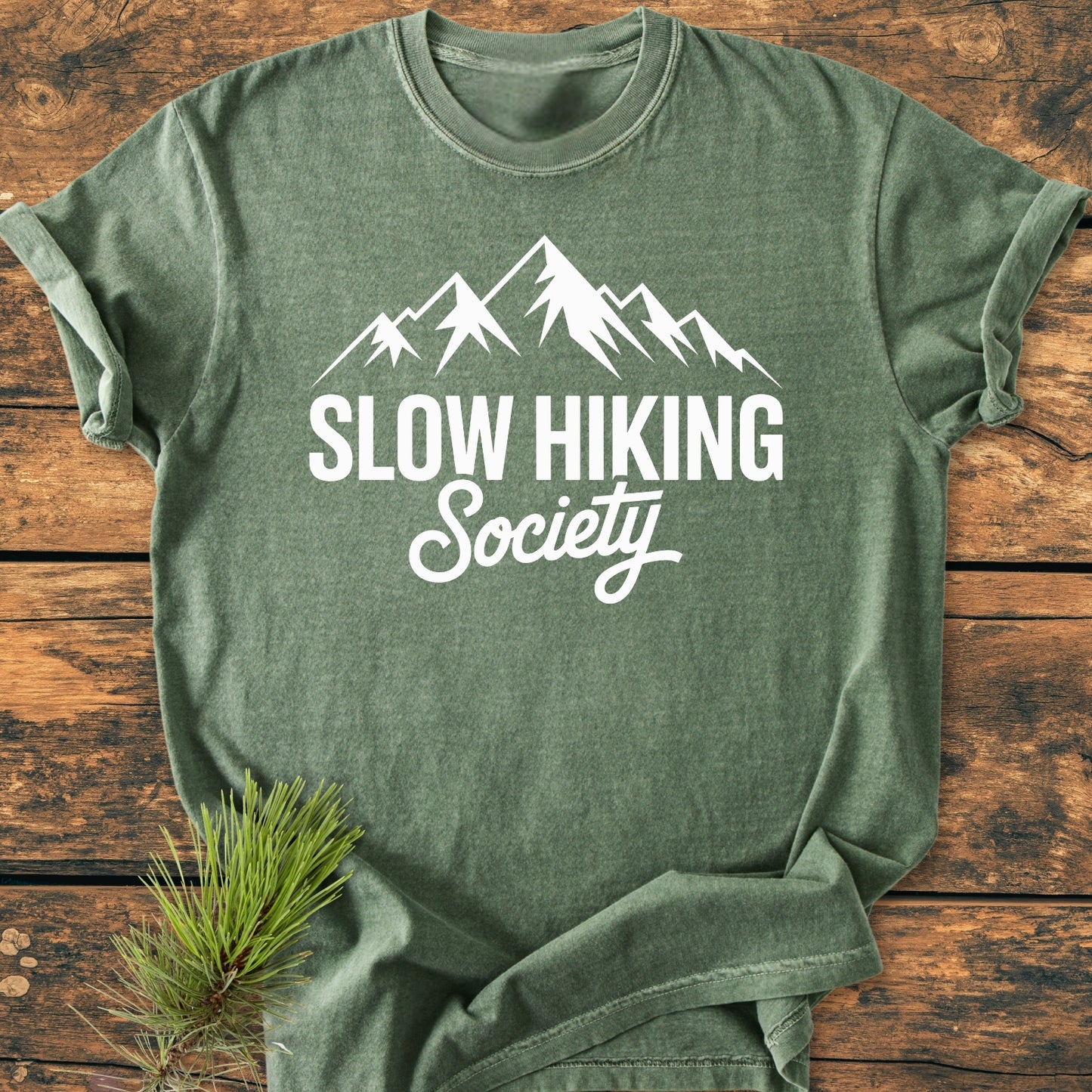 Heather-green Slow Hiking Society - Vintage Wash Tee with stylized mountain graphic, laid flat on wood. Features soft-washed, pre-shrunk fabric and relaxed fit, showcasing durability and comfort.