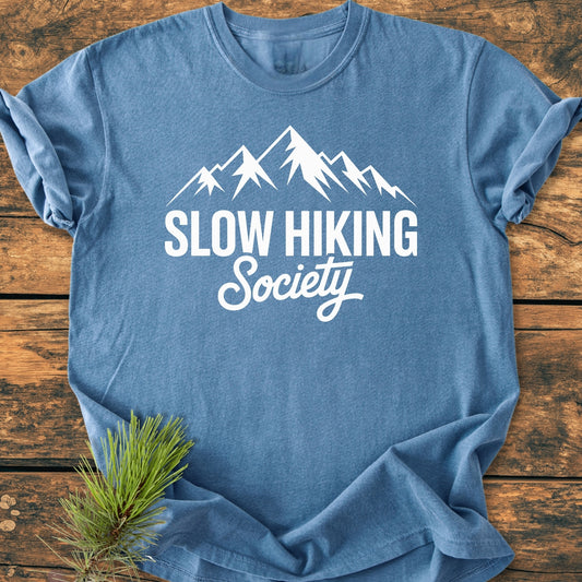 Slow Hiking Society - Vintage Wash Tee featuring a stylized mountain range graphic, laid flat on a wooden surface, with a small pine sprig beside it, highlighting its relaxed fit and durable design.