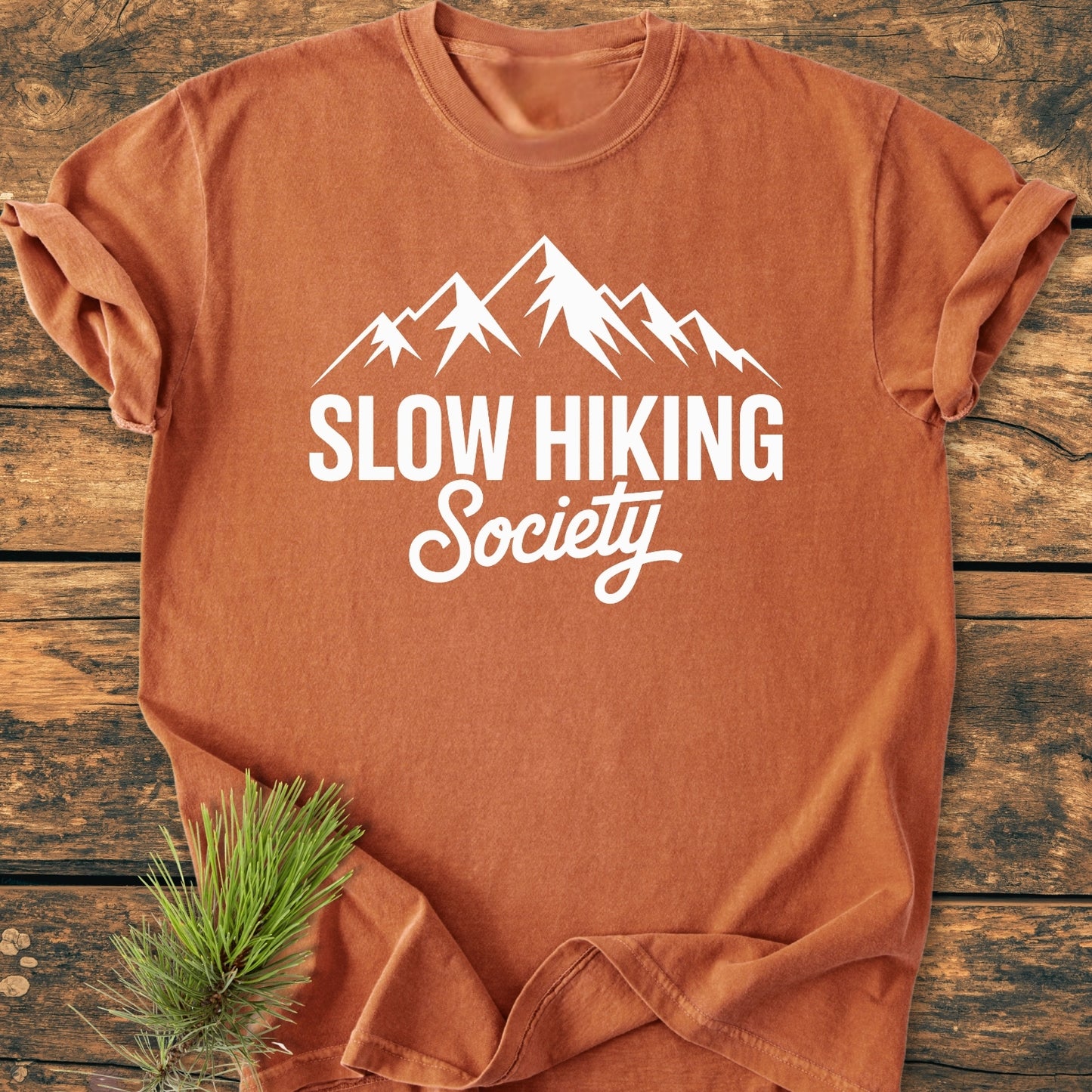 Slow Hiking Society - Vintage Wash Tee: A burnt-orange T-shirt featuring a white mountain graphic and text, laid flat on wood, styled with pine needles. Made of soft, durable garment-dyed cotton.