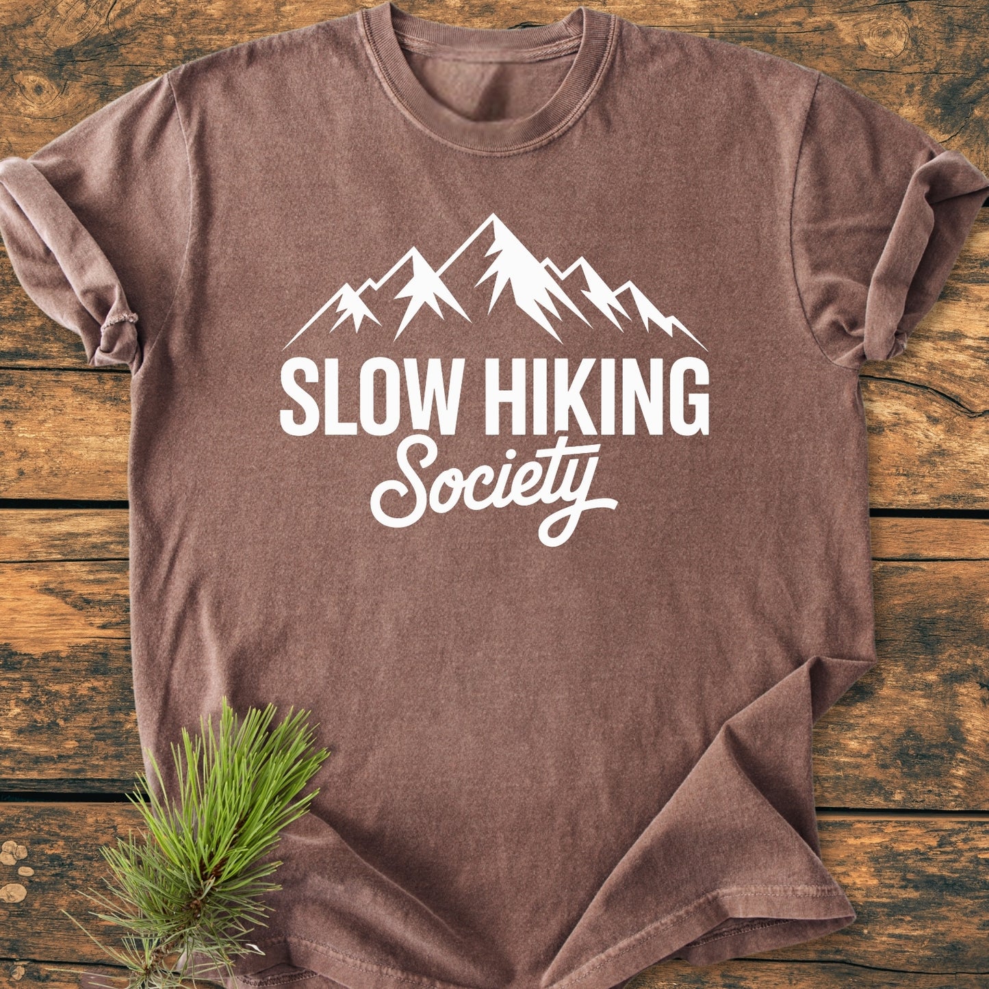 The Slow Hiking Society - Vintage Wash Tee features a mountain graphic and slogan on a soft, brown T-shirt, laid flat on wood, with a pine sprig accenting the design.