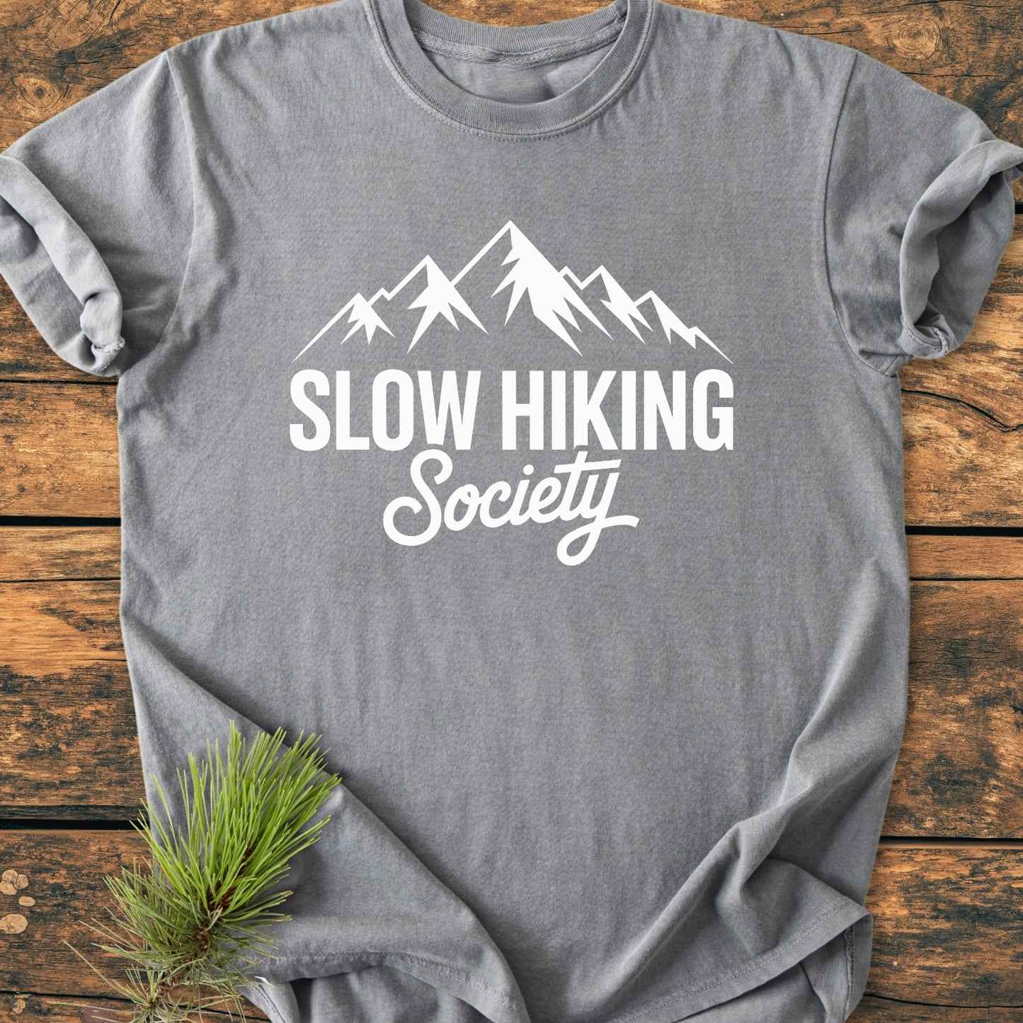 Heather-grey tee with mountain graphic and Slow Hiking Society text, laid on wood. Features soft-washed, garment-dyed fabric with a relaxed fit. Pine sprig accentuates the nature-inspired design.