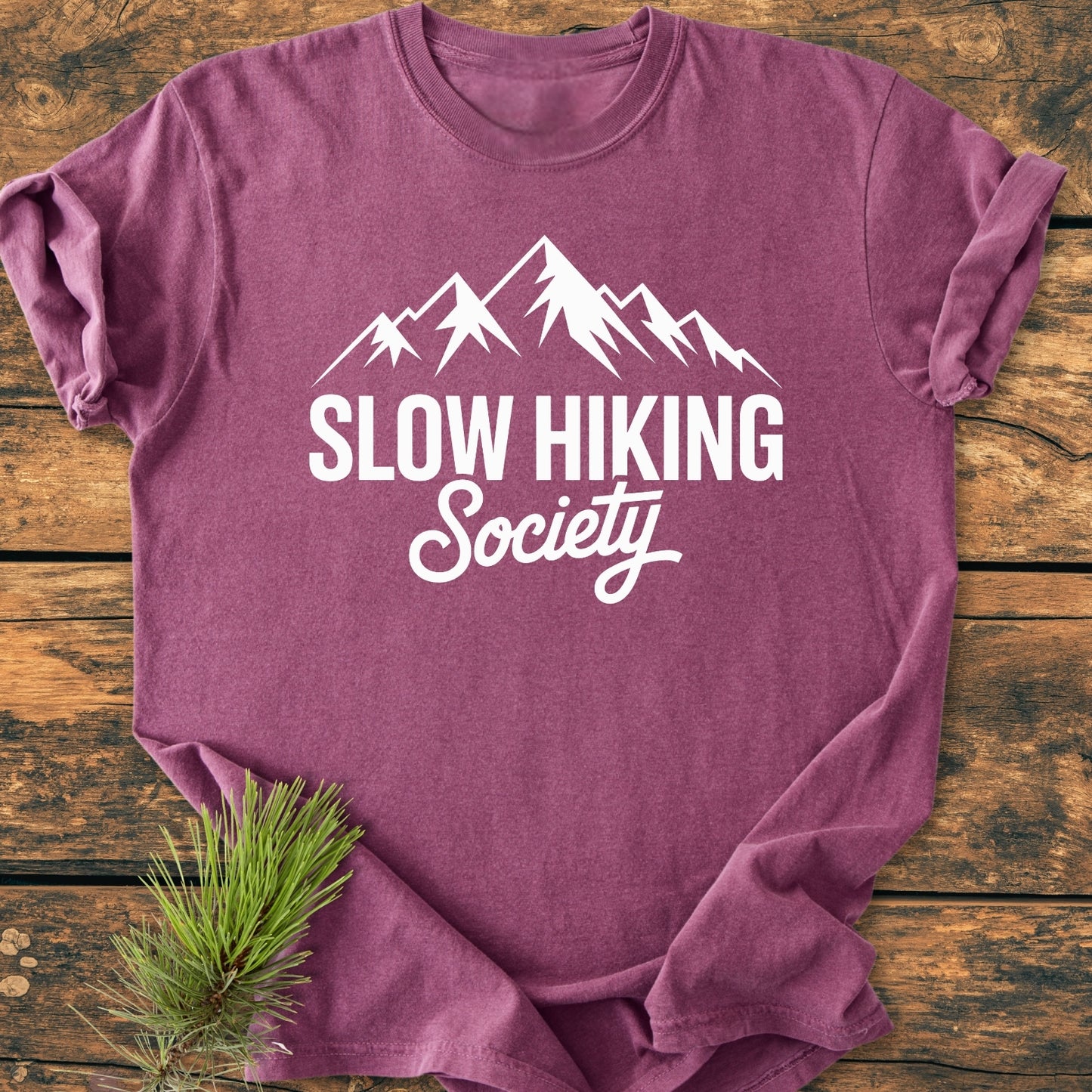 Slow Hiking Society - Vintage Wash Tee features a white mountain graphic and text on a heathered-rose T-shirt, laid flat on wood with a pine sprig, highlighting its relaxed, durable design.