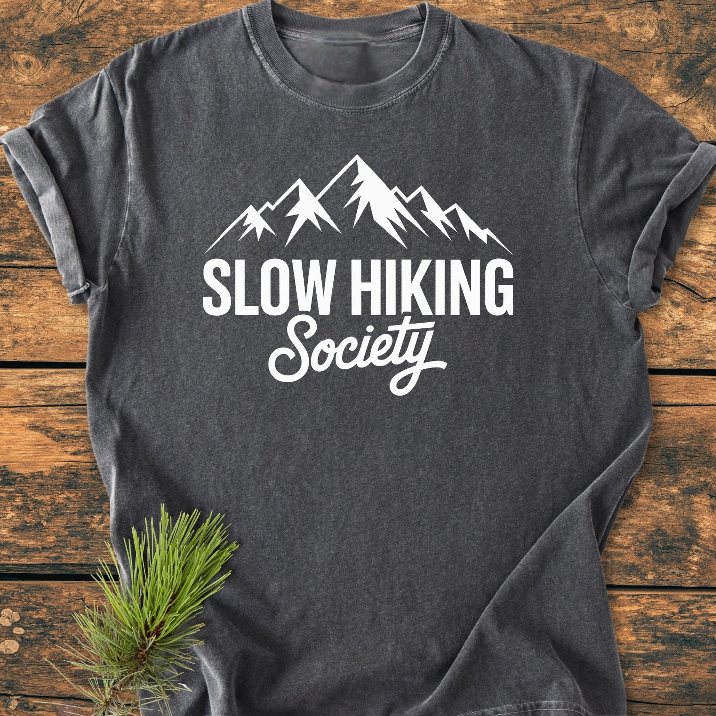 Charcoal-gray Slow Hiking Society - Vintage Wash Tee with white mountain graphic, laid flat on wood surface; features short sleeves and relaxed fit, complemented by a pine sprig in the corner.