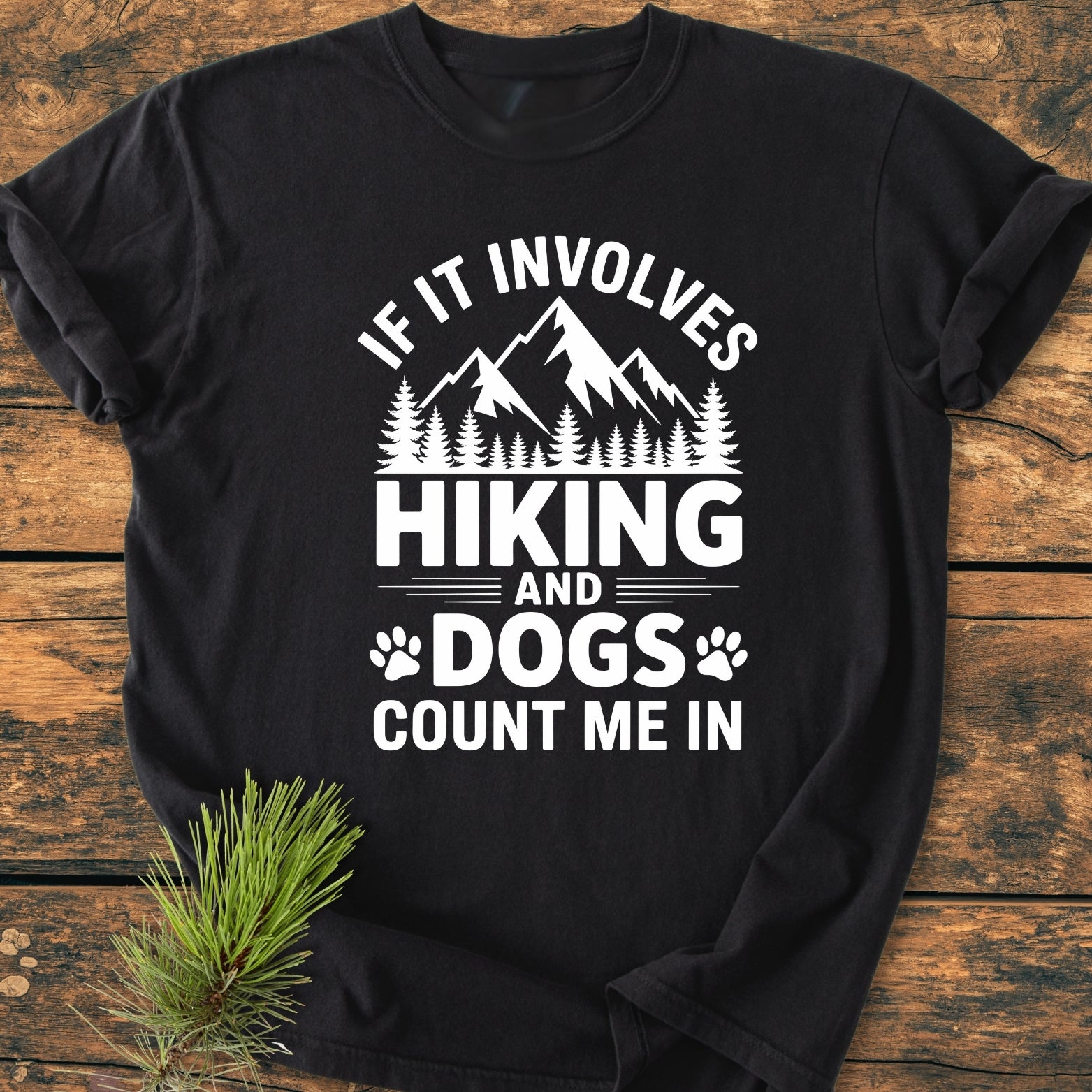 Hiking And Dogs - Vintage Wash Tee, featuring mountain and pine graphics with hiking and dogs text, displayed on a wooden surface with pine branch. Made from soft, durable, pre-shrunk cotton for a cozy fit.