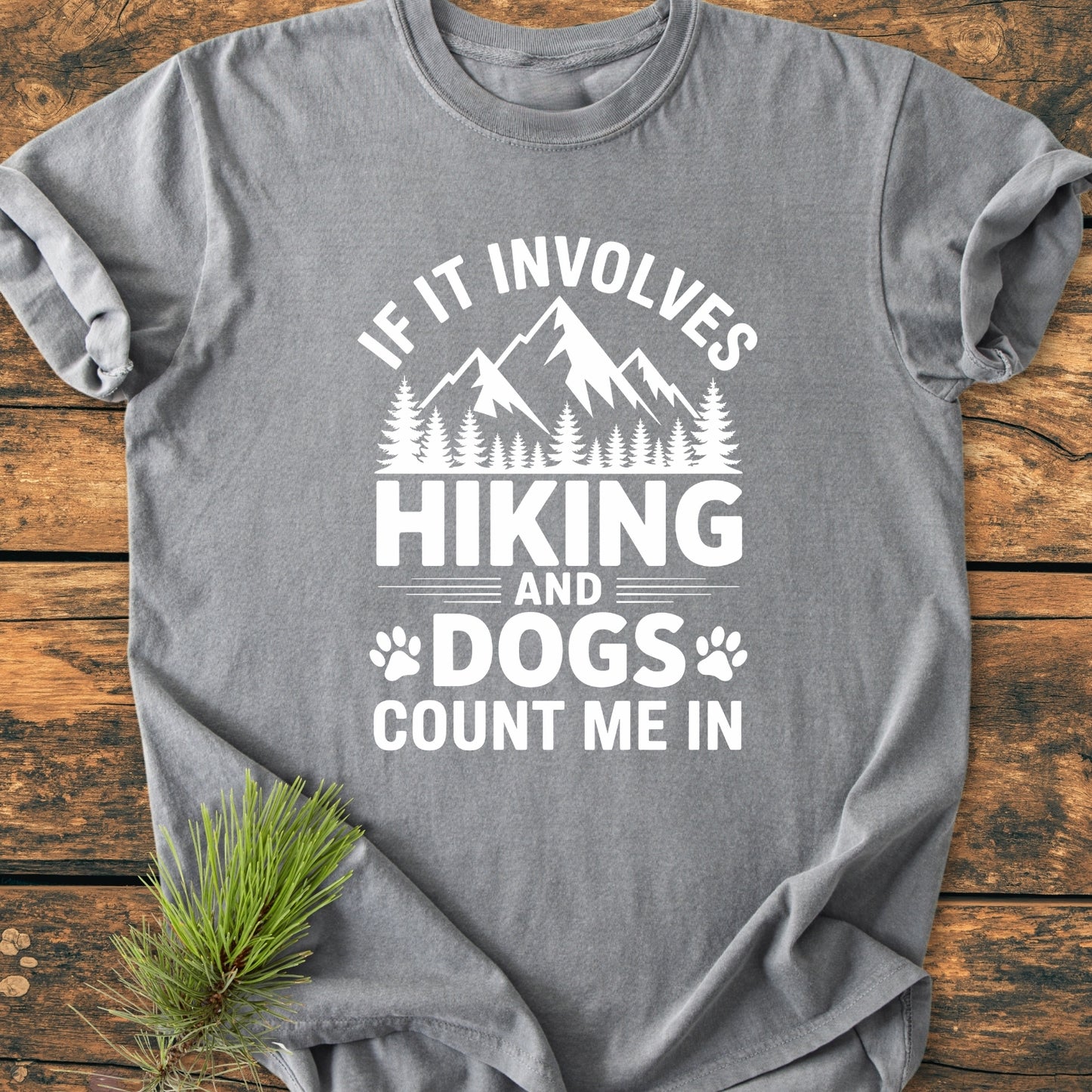 Hiking And Dogs - Vintage Wash Tee featuring a mountain-forest graphic and paw prints on a gray T-shirt, laid flat on wood with pine needles, highlighting an outdoor adventure theme.