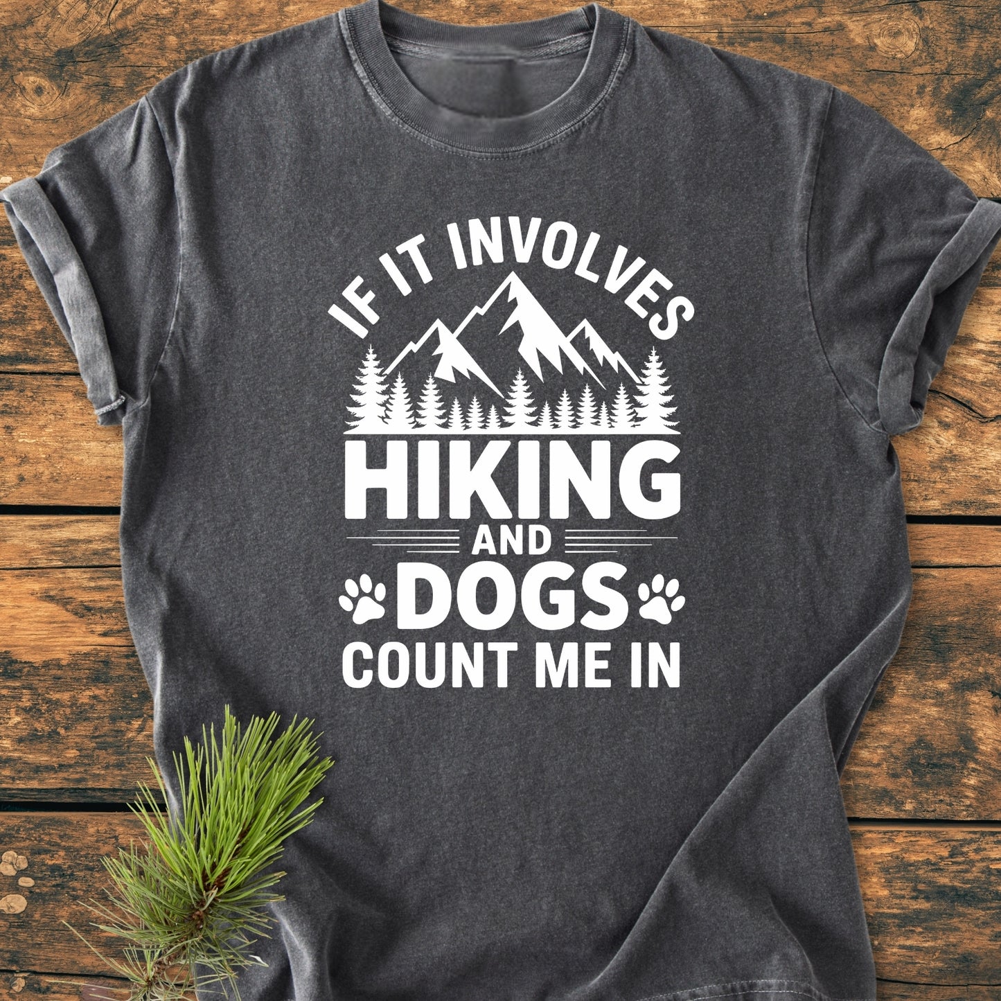 Hiking And Dogs - Vintage Wash Tee features bold mountain and pine tree graphics, with hiking and dog-themed text, laid flat with a pine sprig accent, showcasing its comfortable, relaxed fit.