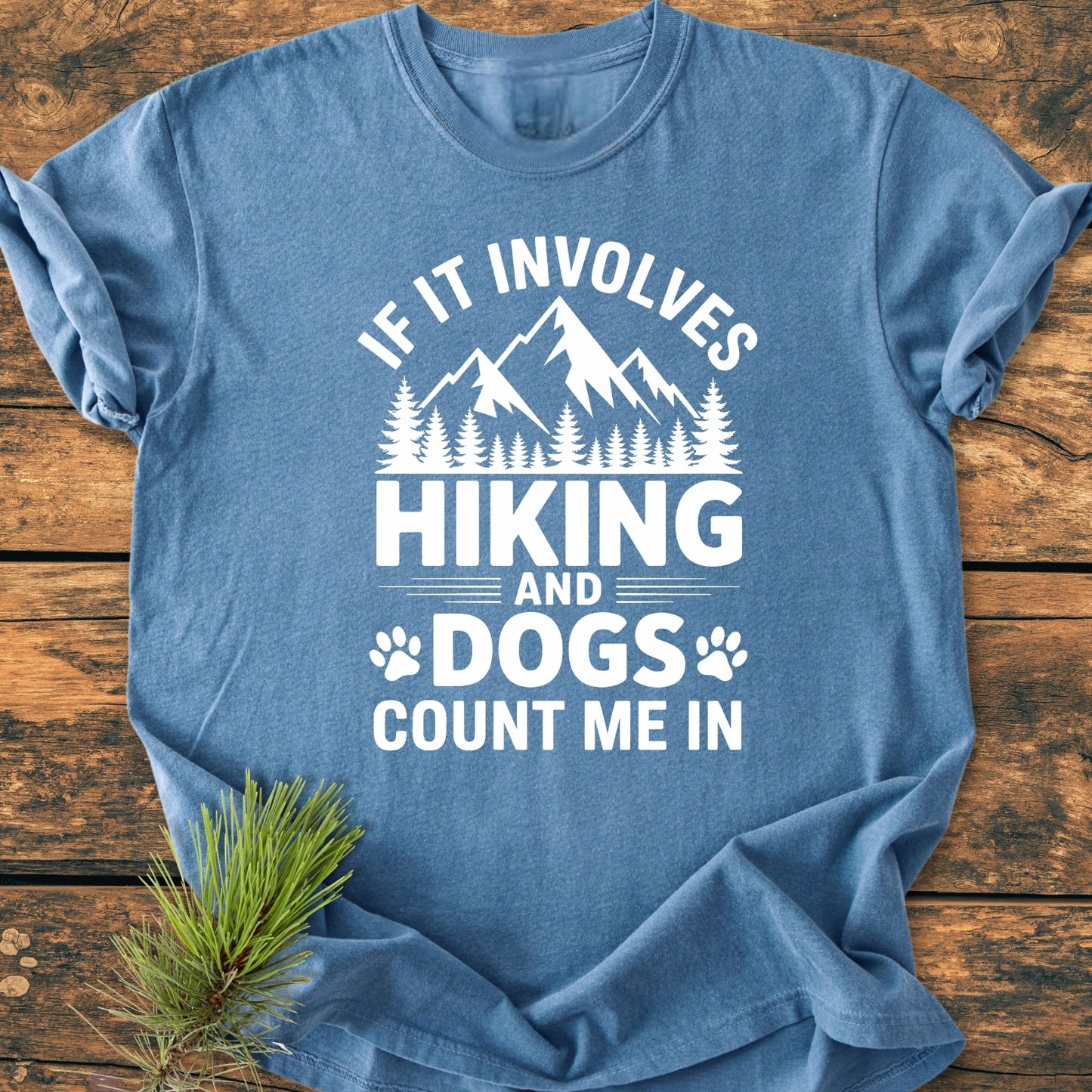 Heather-blue Hiking And Dogs - Vintage Wash Tee on wood, featuring white mountain, tree, and paw print graphic with slogan. Pine sprig accentuates the rustic theme.