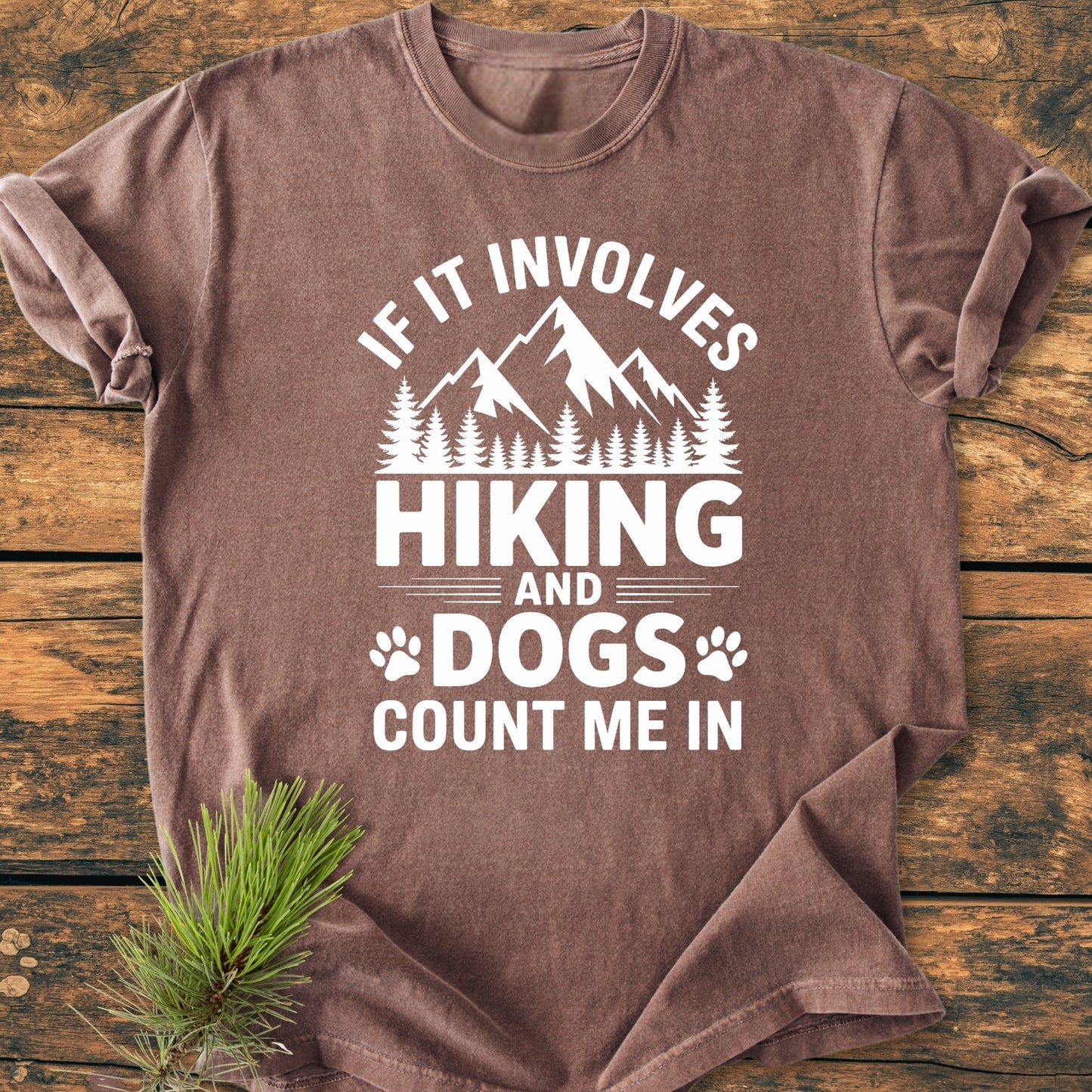Hiking And Dogs - Vintage Wash Tee features a mountain-and-forest graphic with paw prints and slogan on a brown T-shirt, laid on a tabletop with a pine sprig beside it.