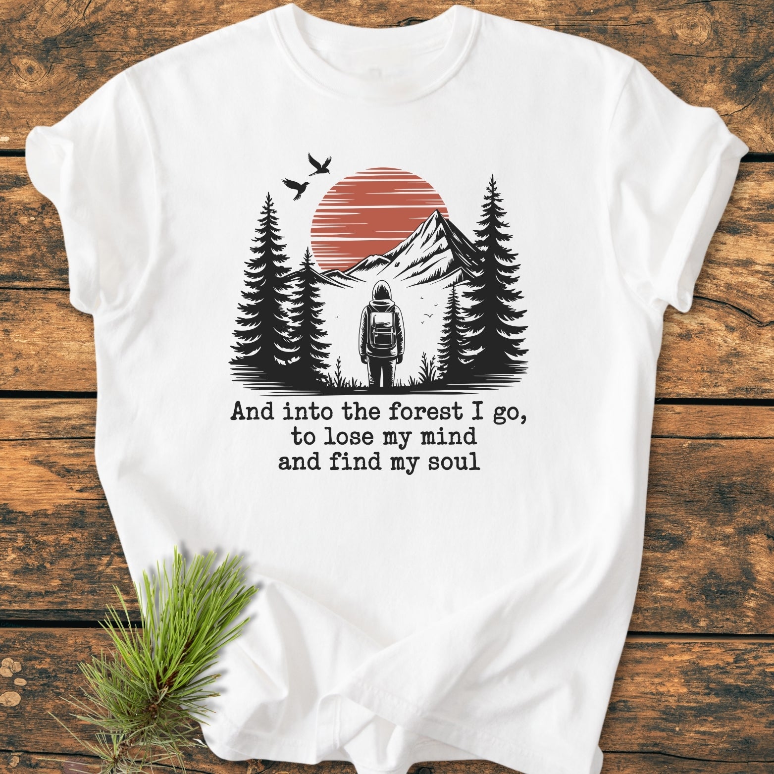 And Into The Forest I Go - Vintage Wash Tee featuring a hiker graphic with pine trees and a red sun, accented by birds and an inspirational quote. Laid on a wooden surface.