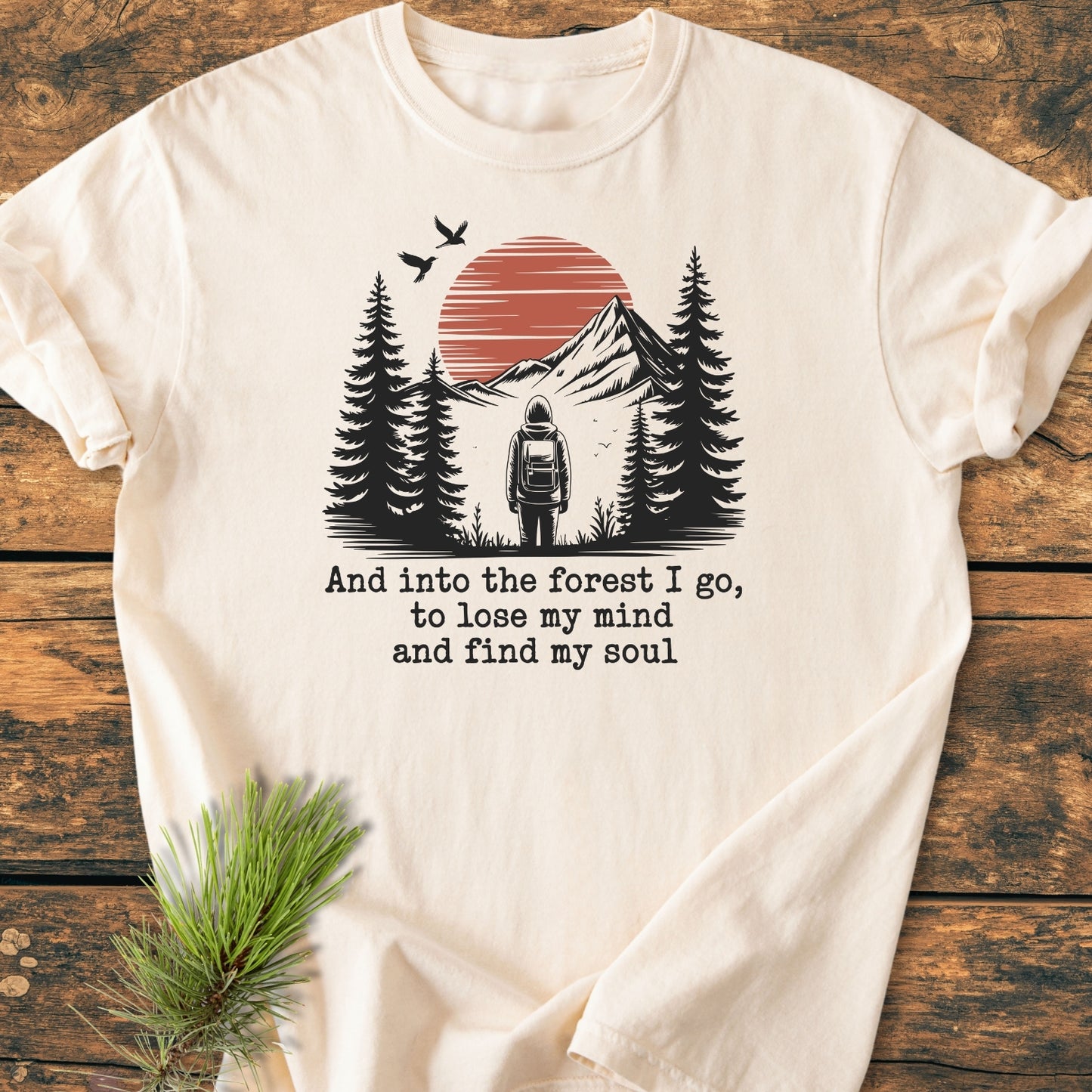 And Into The Forest I Go - Vintage Wash Tee features a stylized nature scene with a backpacked figure among pine trees, a mountain with red sun, and birds, enhancing outdoor adventure vibes.