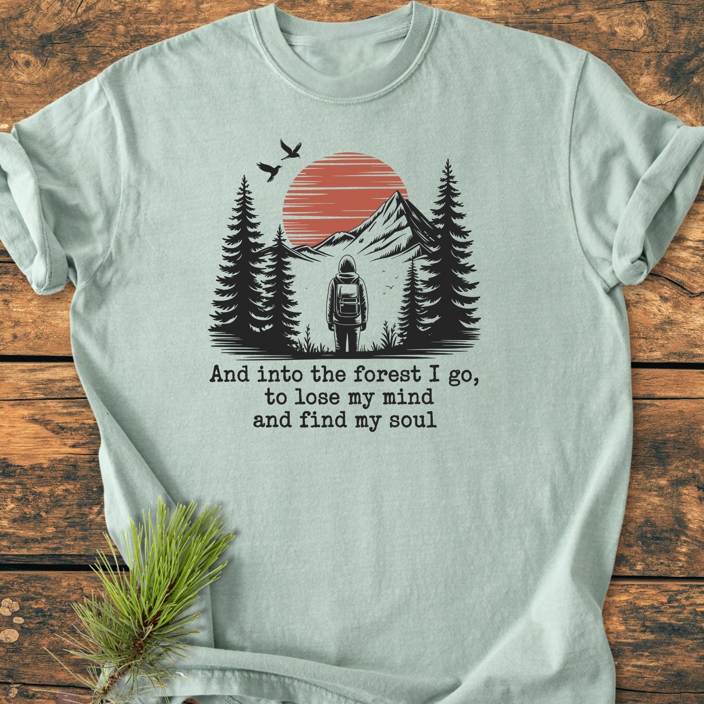 And Into The Forest I Go - Vintage Wash Tee featuring a hiker graphic with mountains and birds, laid flat on wood. Includes poetic text, crafted from garment-dyed, soft-washed cotton for comfort and durability.