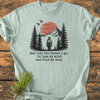 And Into The Forest I Go - Vintage Wash Tee featuring a hiker graphic with mountains and birds, laid flat on wood. Includes poetic text, crafted from garment-dyed, soft-washed cotton for comfort and durability.