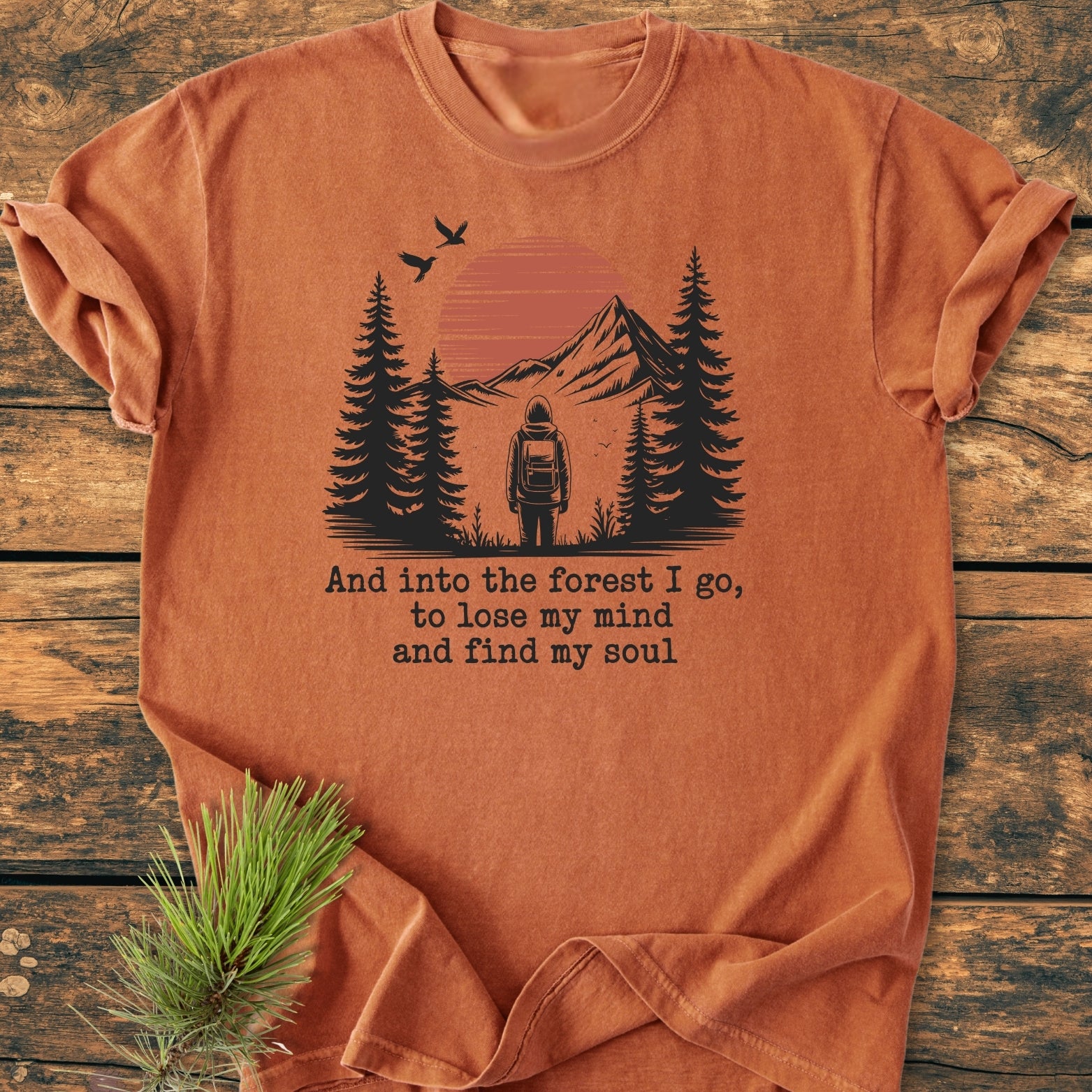 And Into The Forest I Go - Vintage Wash Tee, featuring a serene outdoor scene with a hiker, pine trees, mountains, setting sun, and birds, printed on a soft-rust, folded T-shirt.