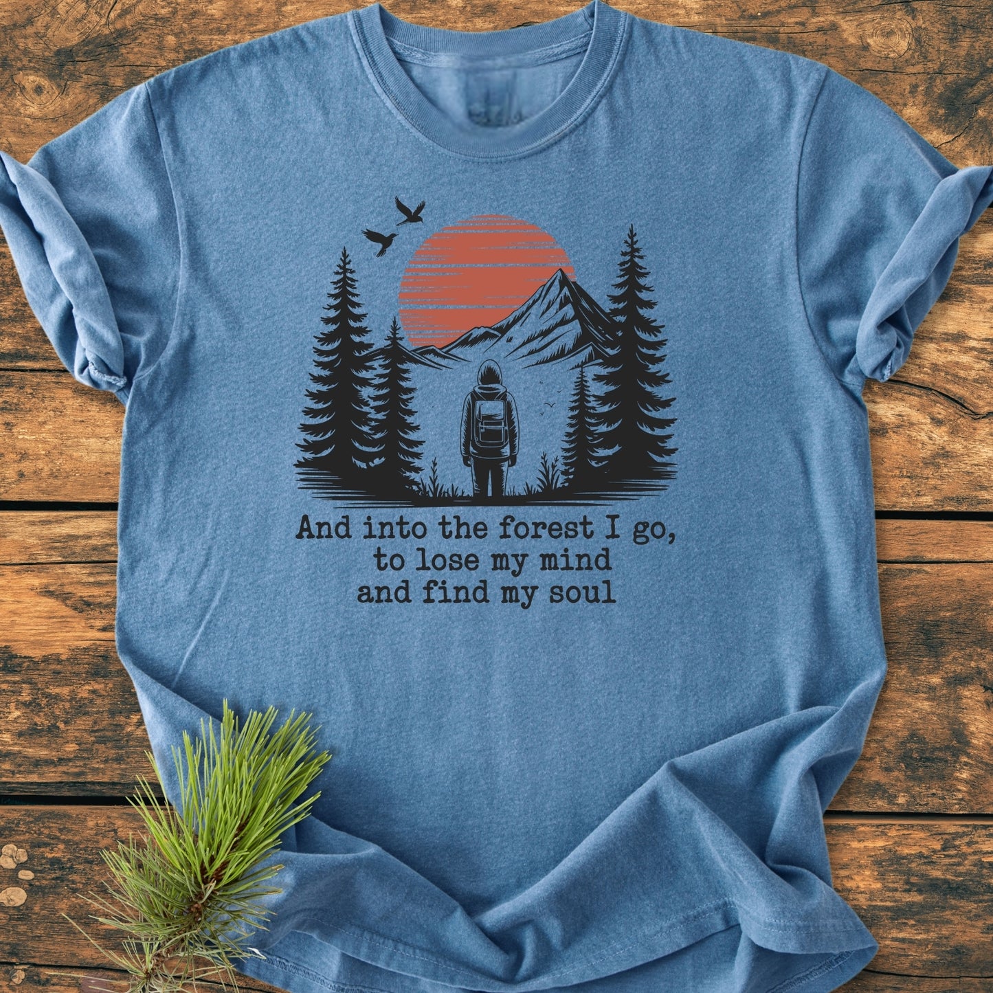 And Into The Forest I Go - Vintage Wash Tee features a hiker silhouette among pine trees, with a mountain range and stylized sun behind, on soft, relaxed-fit fabric.