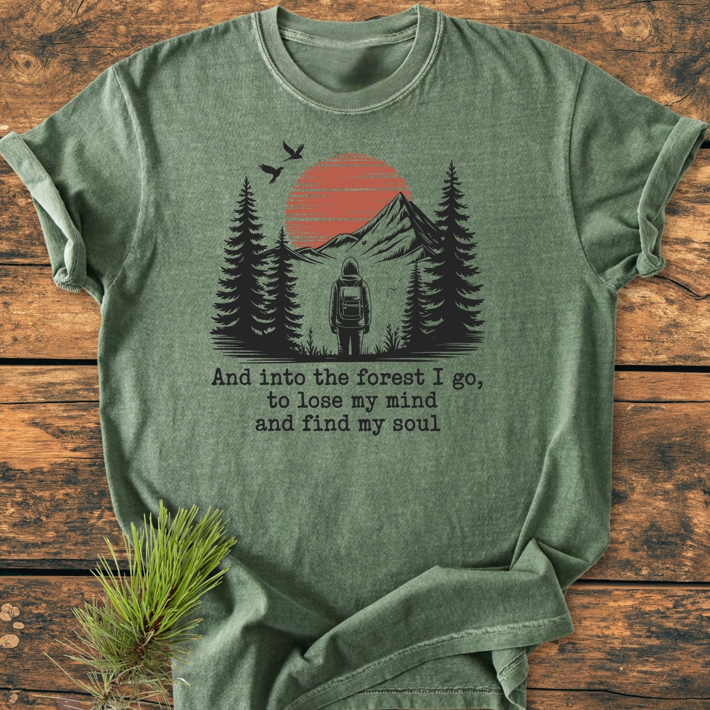 And Into The Forest I Go - Vintage Wash Tee features a stylized outdoor scene with a backpacker, pine trees, and mountains. The soft olive-green fabric, made from 100% ring-spun cotton, ensures comfort.