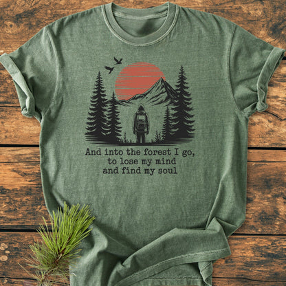 And Into The Forest I Go - Vintage Wash Tee features a stylized outdoor scene with a backpacker, pine trees, and mountains. The soft olive-green fabric, made from 100% ring-spun cotton, ensures comfort.