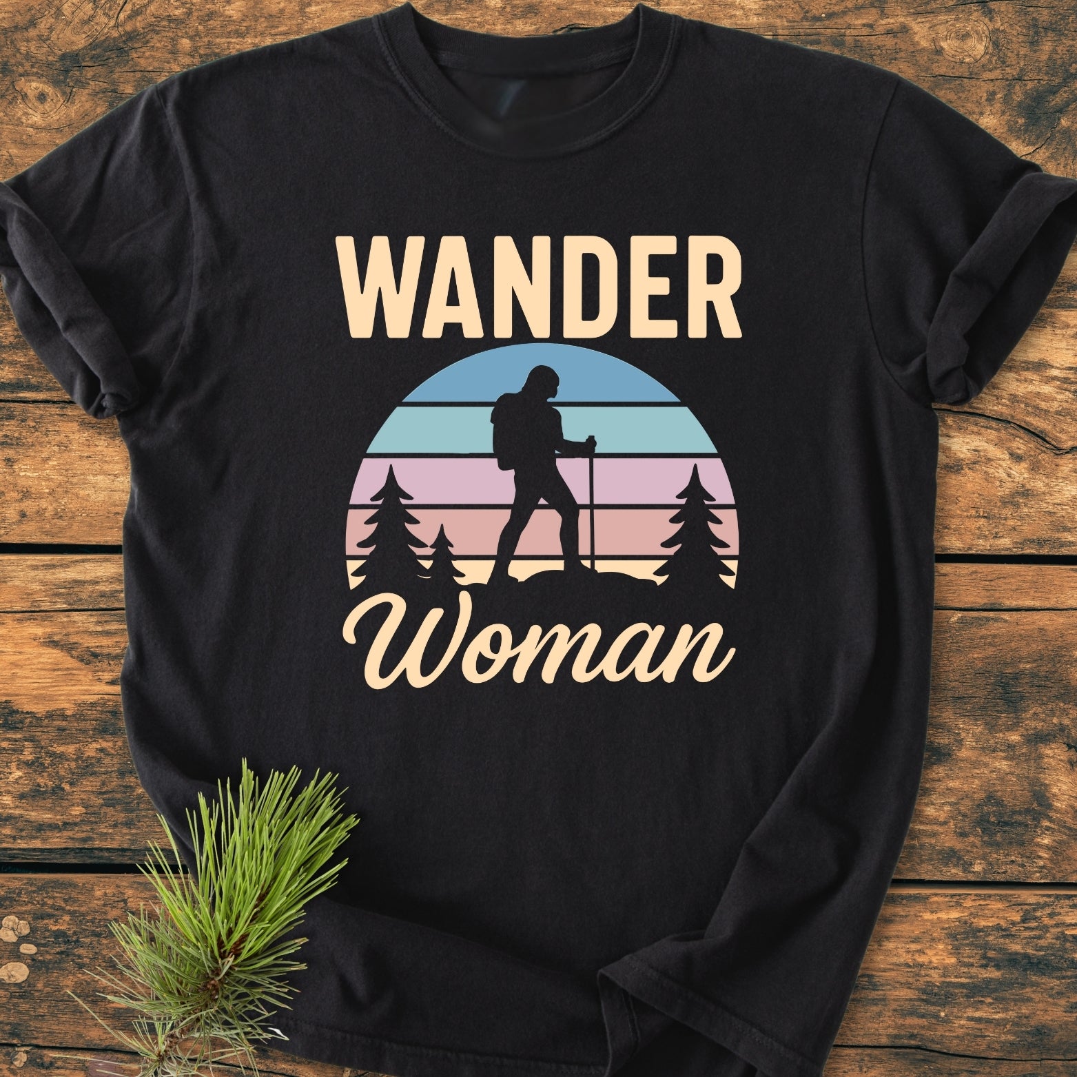 Wander Woman Retro - Vintage Wash Tee featuring a woman hiker graphic with pine trees and sunset, laid on a wooden surface. Includes WANDER text above and Woman script below.