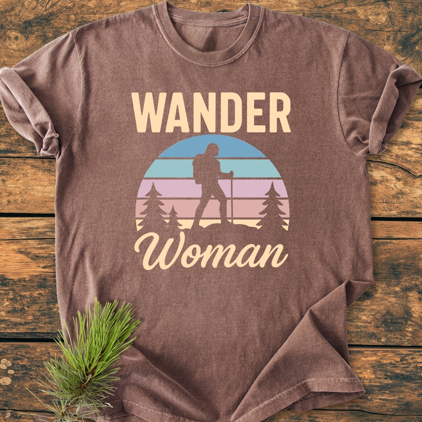 Wander Woman Retro - Vintage Wash Tee on wooden surface, featuring a hiker graphic with 'WANDER' and 'Woman' text. Includes evergreen sprig. Made from soft, durable, pre-shrunk 100% ring-spun cotton.