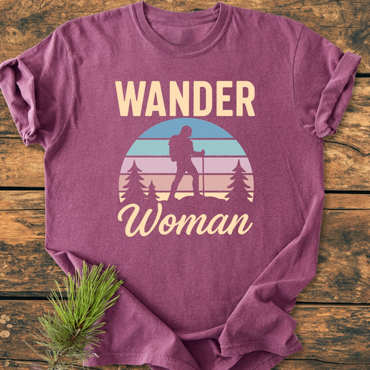 Wander Woman Retro - Vintage Wash Tee features a stylized sunset with a hiker silhouette among pine trees, printed on a heathered plum T-shirt, displayed with a pine sprig.