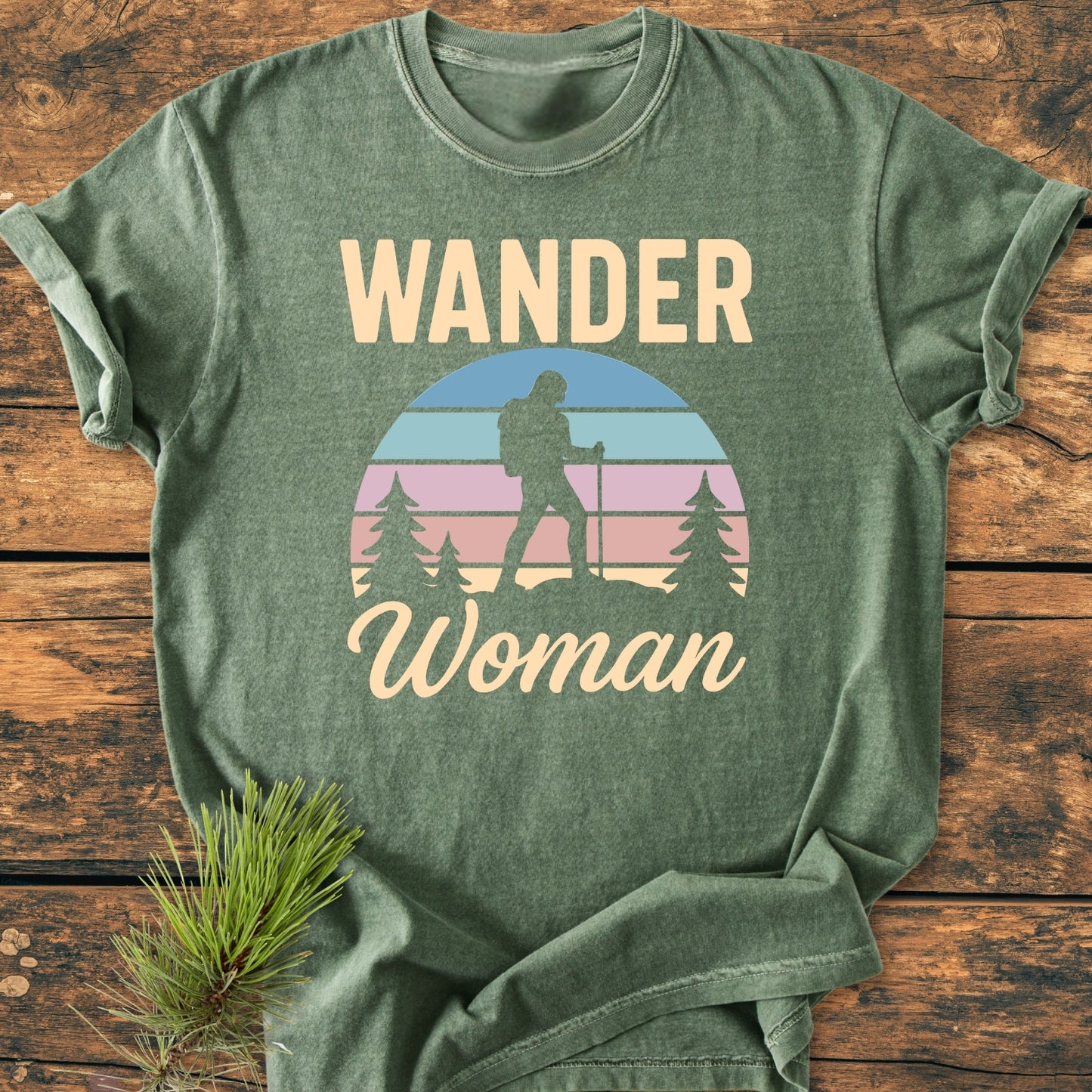 Heather-green Wander Woman Retro - Vintage Wash Tee on wooden surface, featuring WANDER Woman text and silhouette of a woman hiking with pine trees in pastel-striped circular graphic. Pine branch accent nearby.