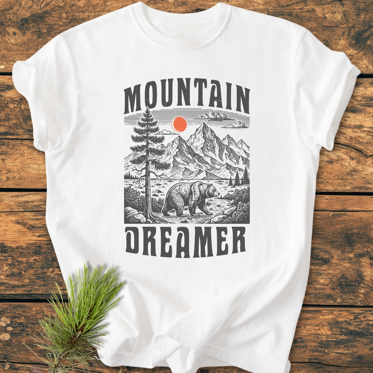 Mountain Dreamer - Vintage Wash Tee featuring a stylized mountain scene with peaks, a bear, and sun. Laid flat on wood, includes a nearby pine sprig, highlighting its nature-inspired design.