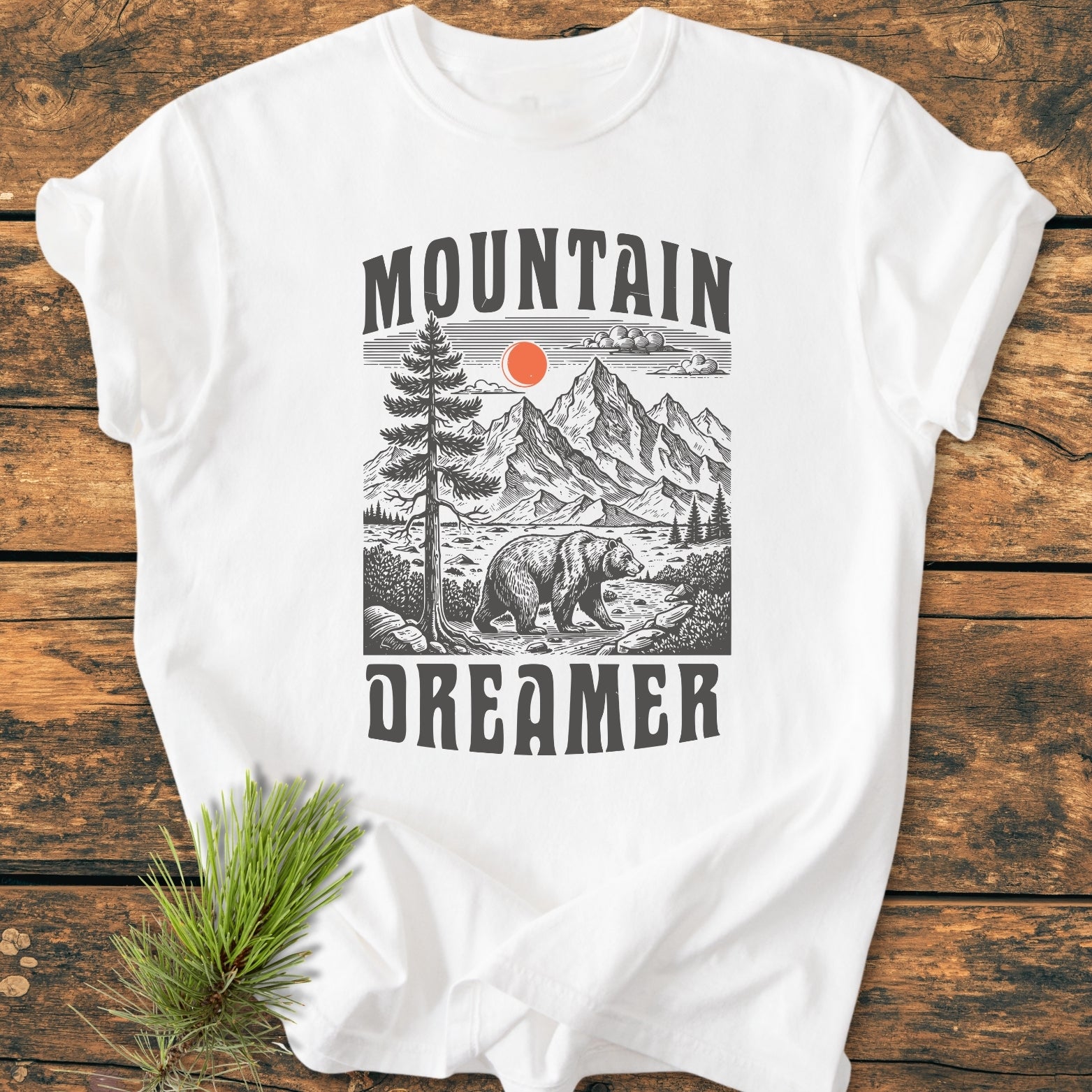 Mountain Dreamer - Vintage Wash Tee featuring a stylized mountain scene with peaks, a bear, and sun. Laid flat on wood, includes a nearby pine sprig, highlighting its nature-inspired design.