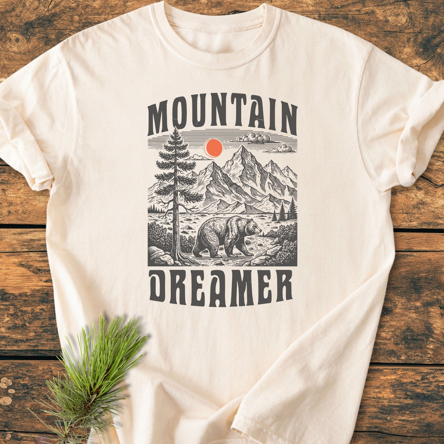 Mountain Dreamer - Vintage Wash Tee features a stylized mountain scene with jagged peaks, a bear, and an orange sun, framed by MOUNTAIN and DREAMER, laid on a wooden surface.